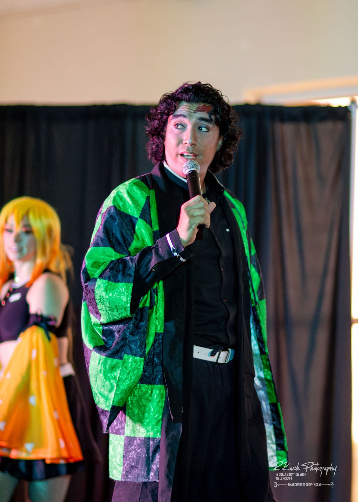 A performer in a green and black patchwork jacket holds a microphone while standing on stage with dramatic lighting.