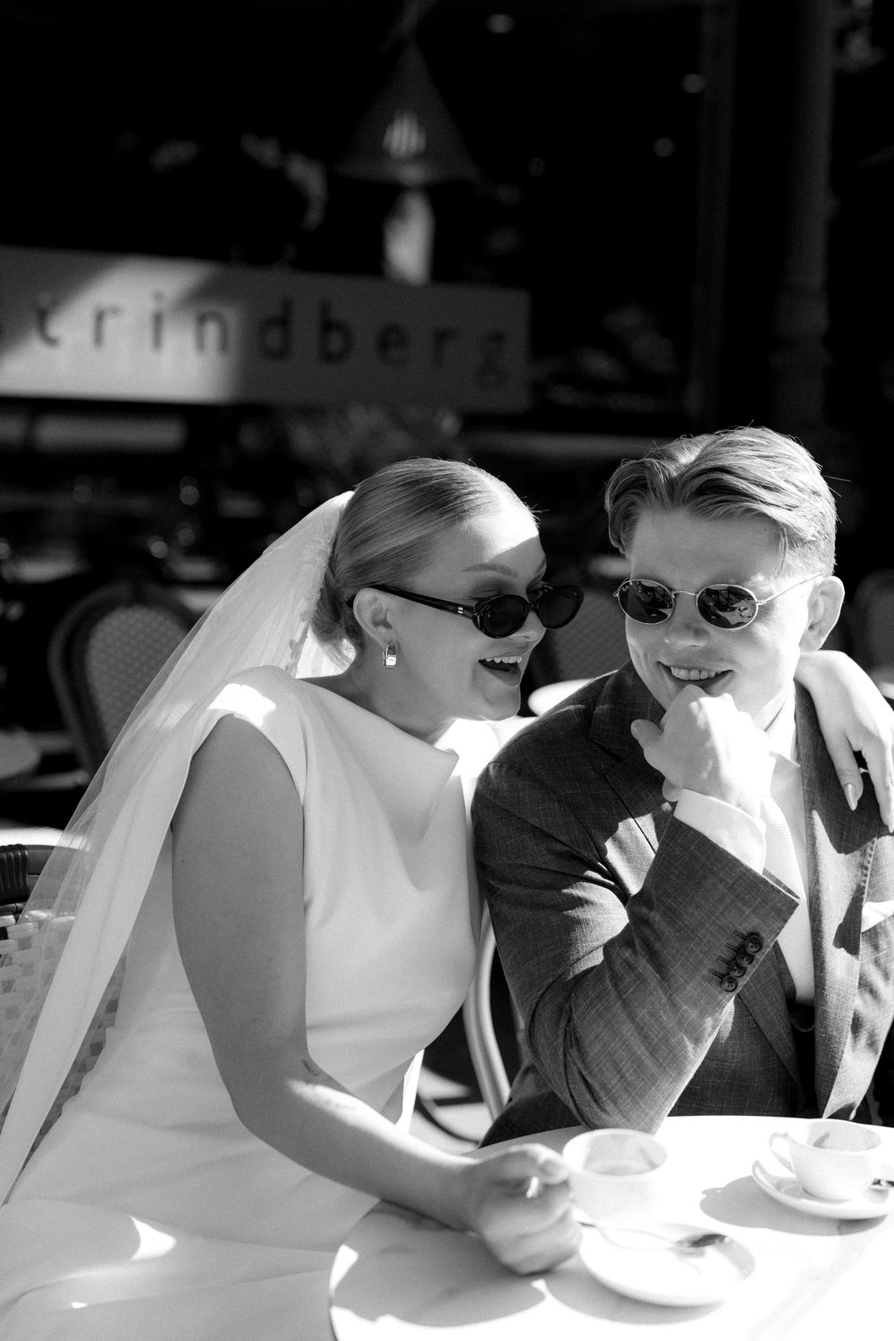 A stylish wedding portrait of bride and groom wearing sunglasses sharing a candid moment at an outdoor cafe table in Helsinki, Finland.