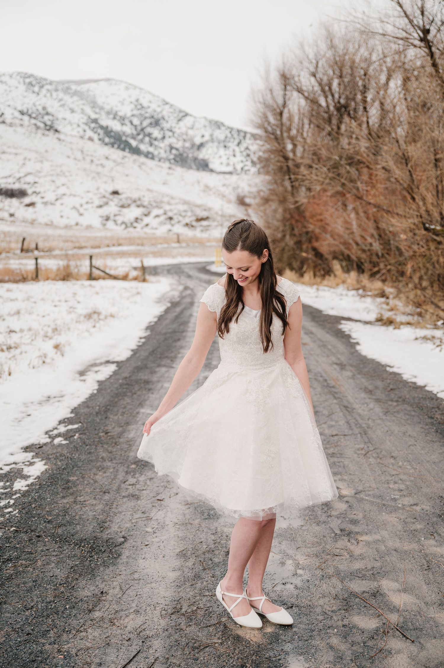 A person in a white dress and ballet flats twirls on a snowy mountain road with bare trees in the background.