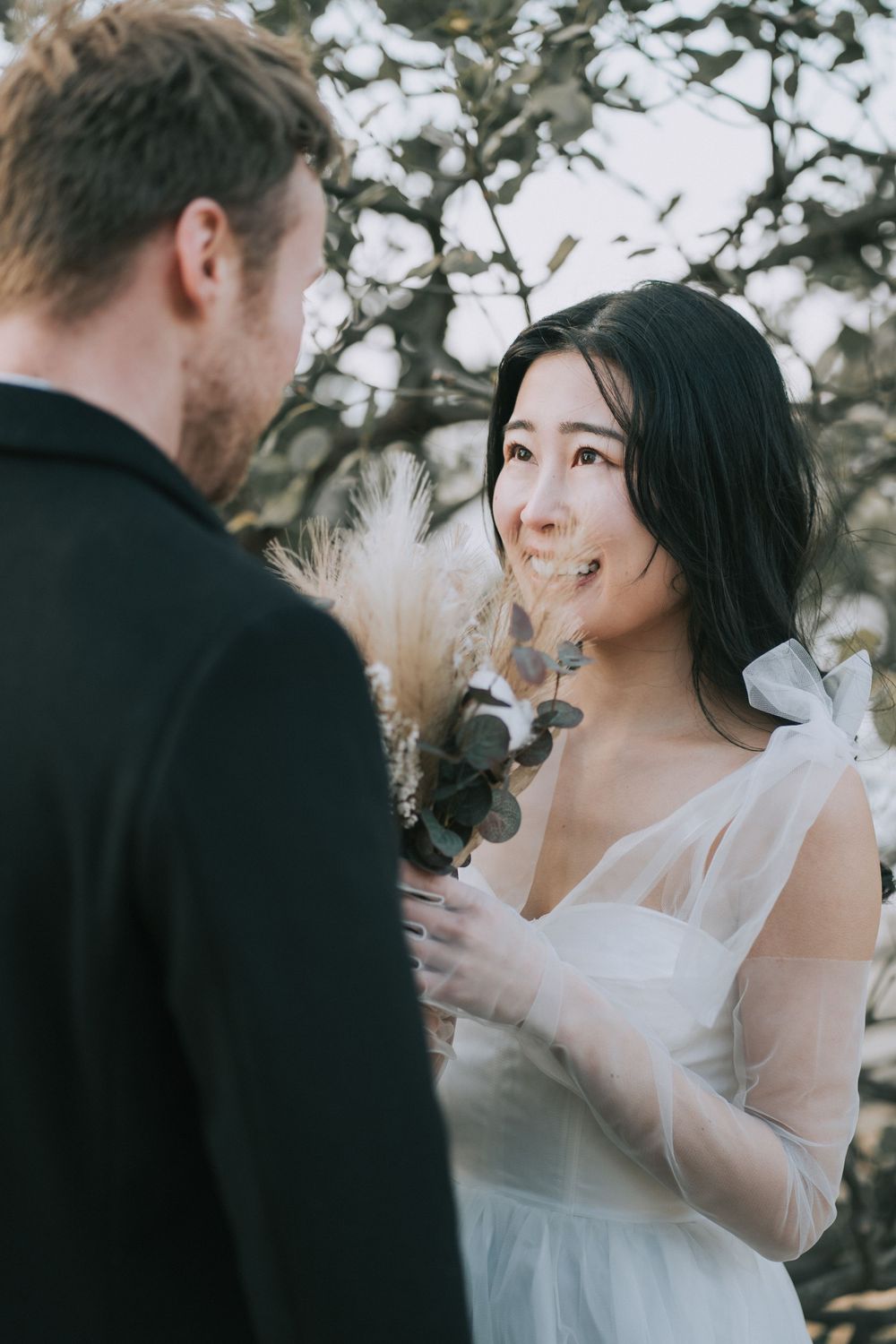 Bride wearing a wedding dress, holding a bouquet up to her face, smiling at the groom