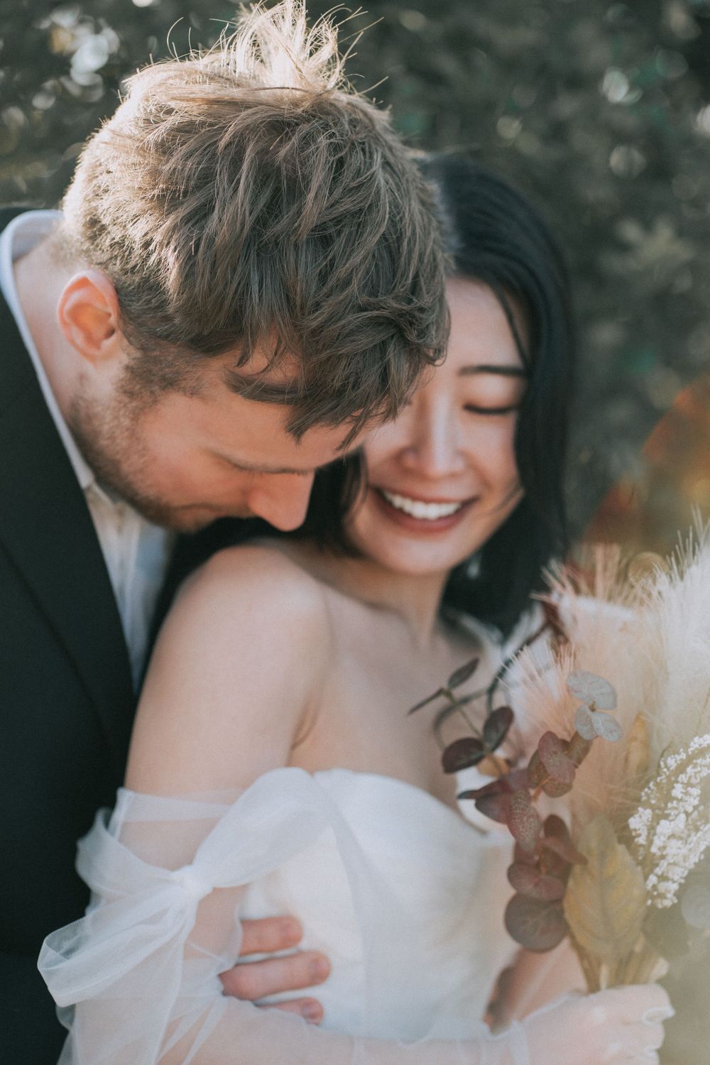 A man is kissing his newly wife on her shoulder, hugging her from behind