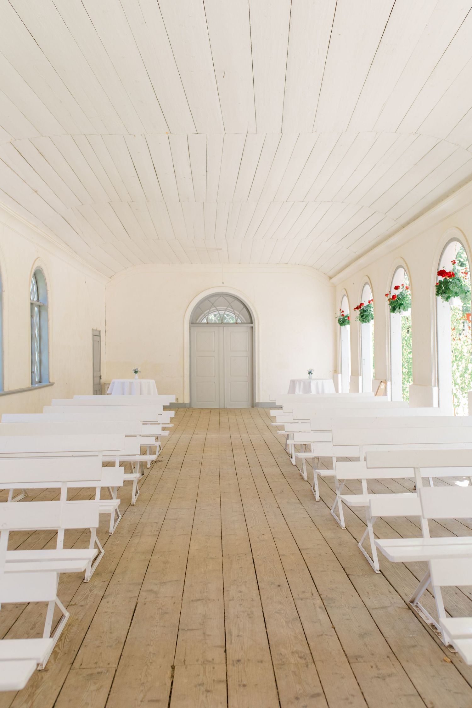 Interior view of a bright white chapel with wooden floors, arched windows and white chairs set up for a ceremony.