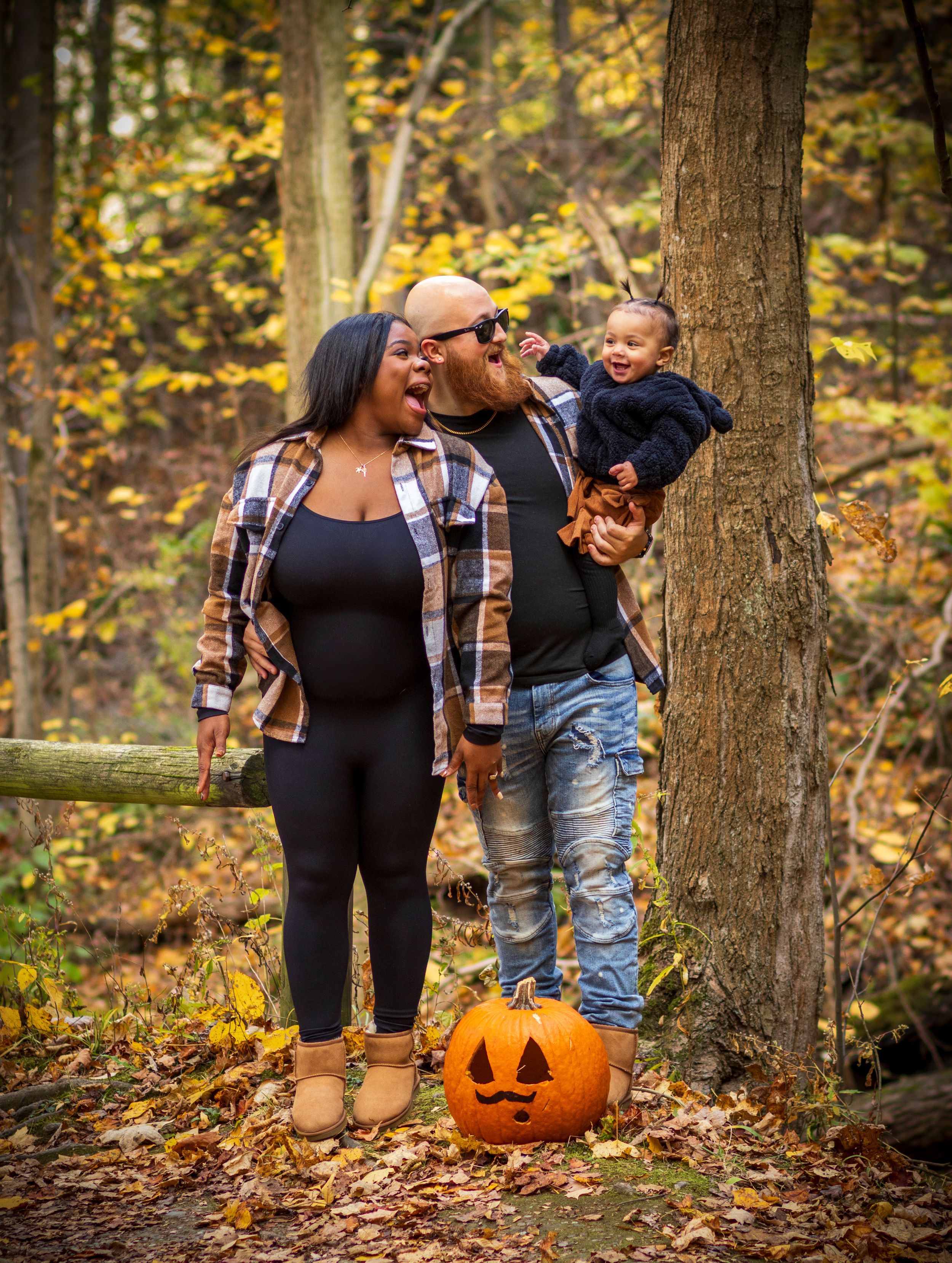 A happy family of three in a forest, with the father holding their baby and a carved pumpkin in front, showcasing a fall family photoshoot