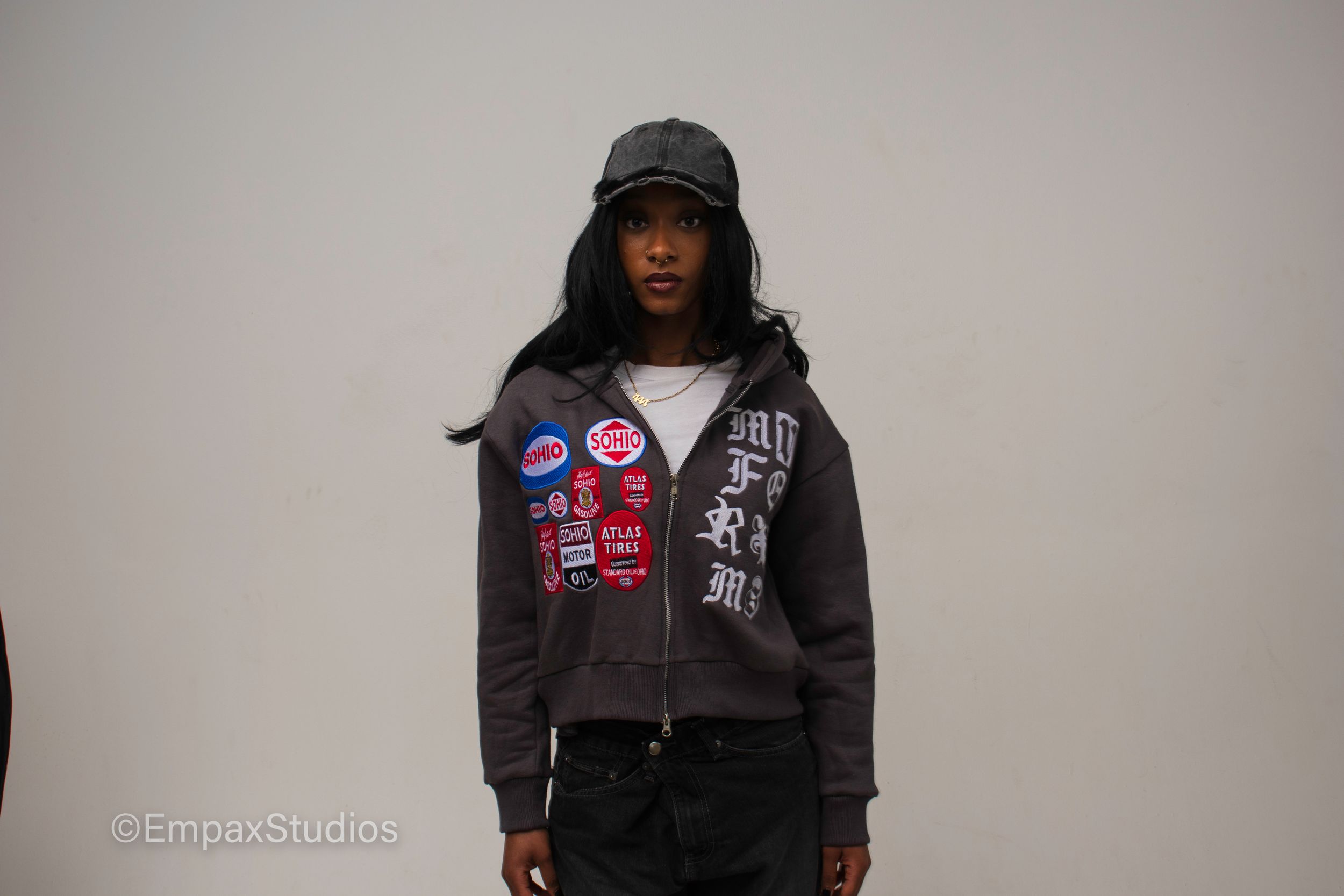 A model wearing a graphic hoodie and cap poses against a neutral background, showcasing a fashion shoot for a streetwear brand.