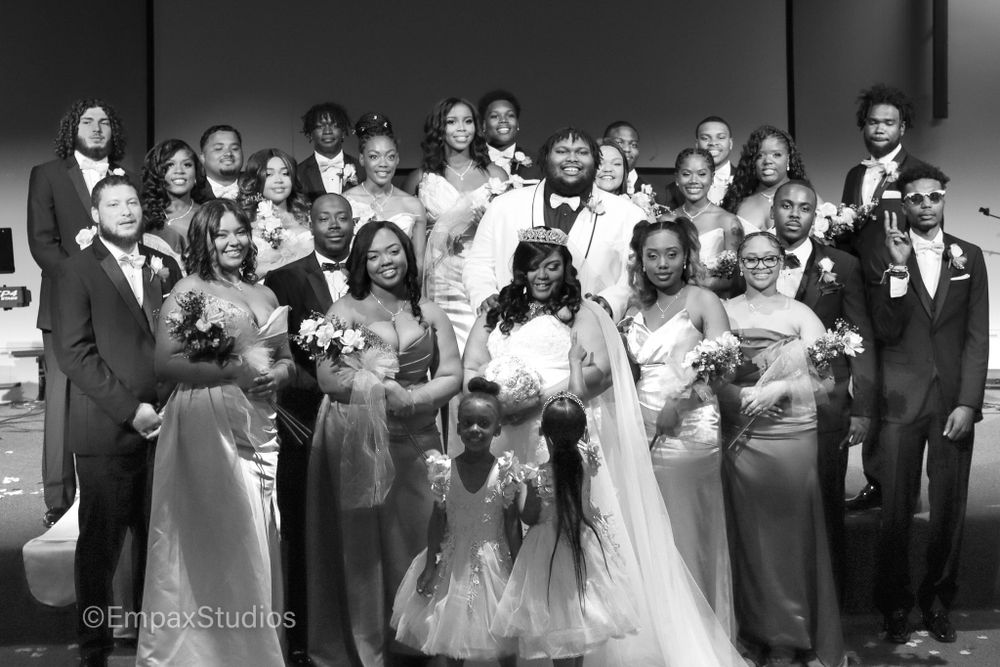 A black-and-white group wedding photo featuring the bride, groom, bridal party smiling together after the ceremony.