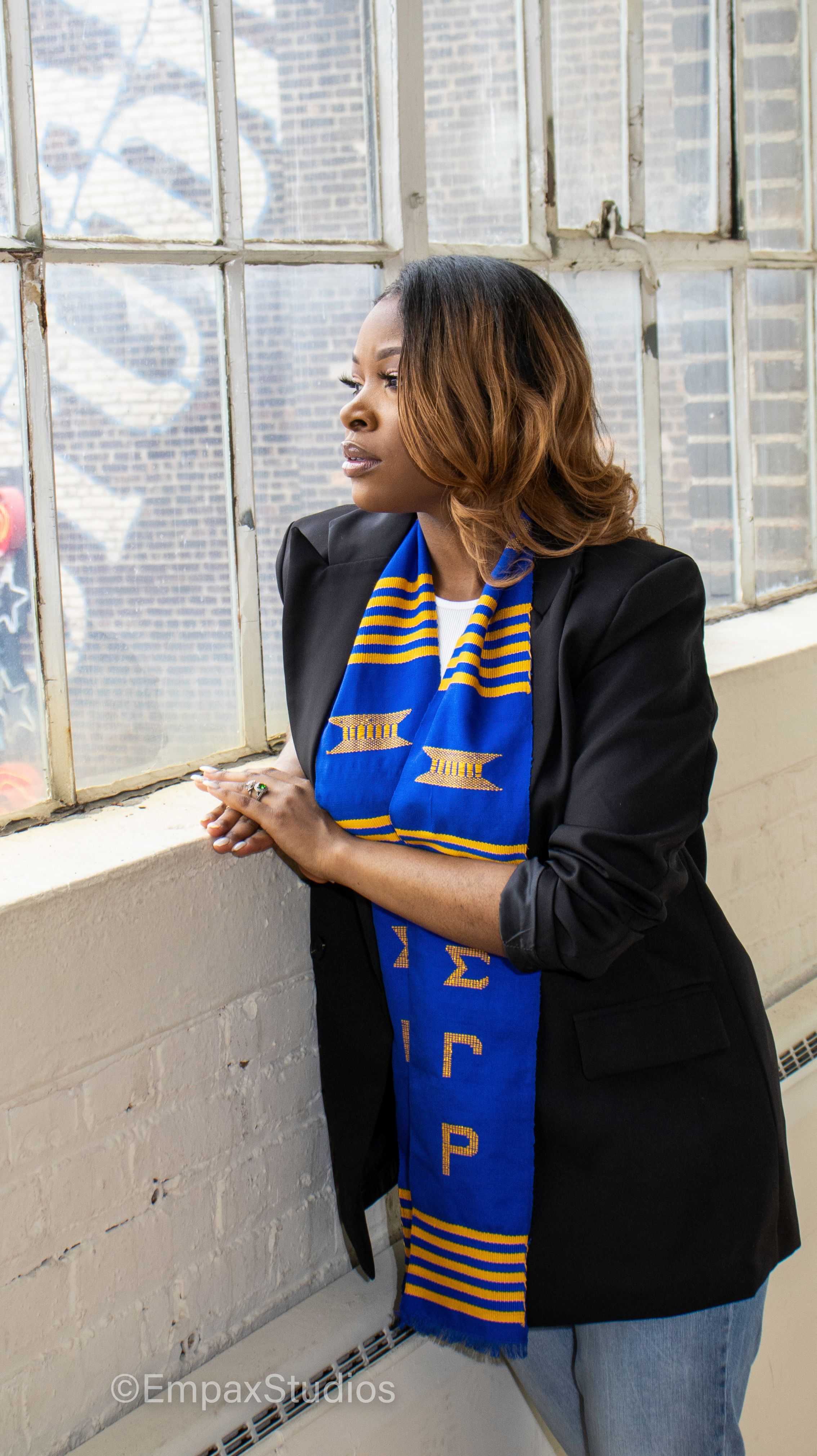 A graduate wearing a blue and gold sorority stole poses by a window during a graduation photoshoot, looking confidently into the distance.