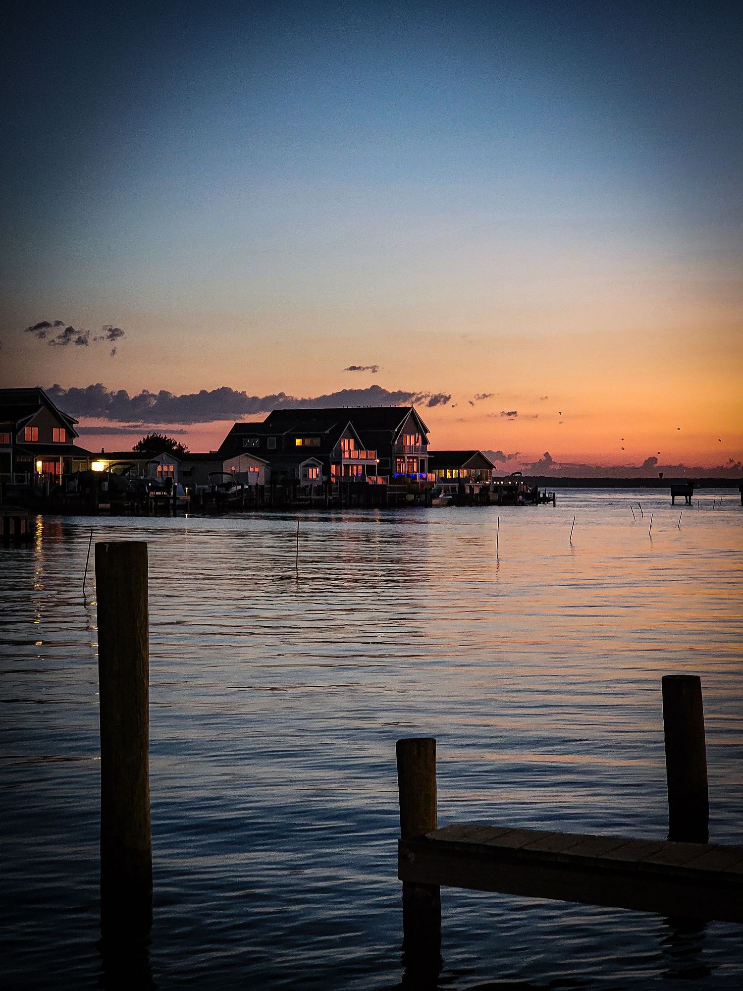 Waterfront sunset with silhouetted beach houses and wooden pier posts reflecting on calm water.