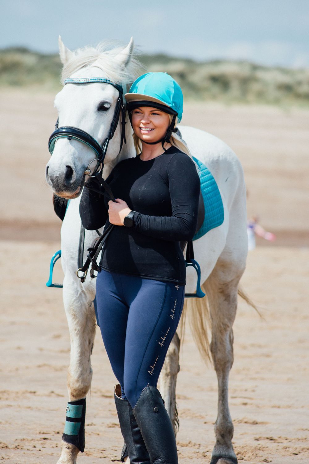 A white horse wearing turquoise tack equipment stands on a sandy beach.