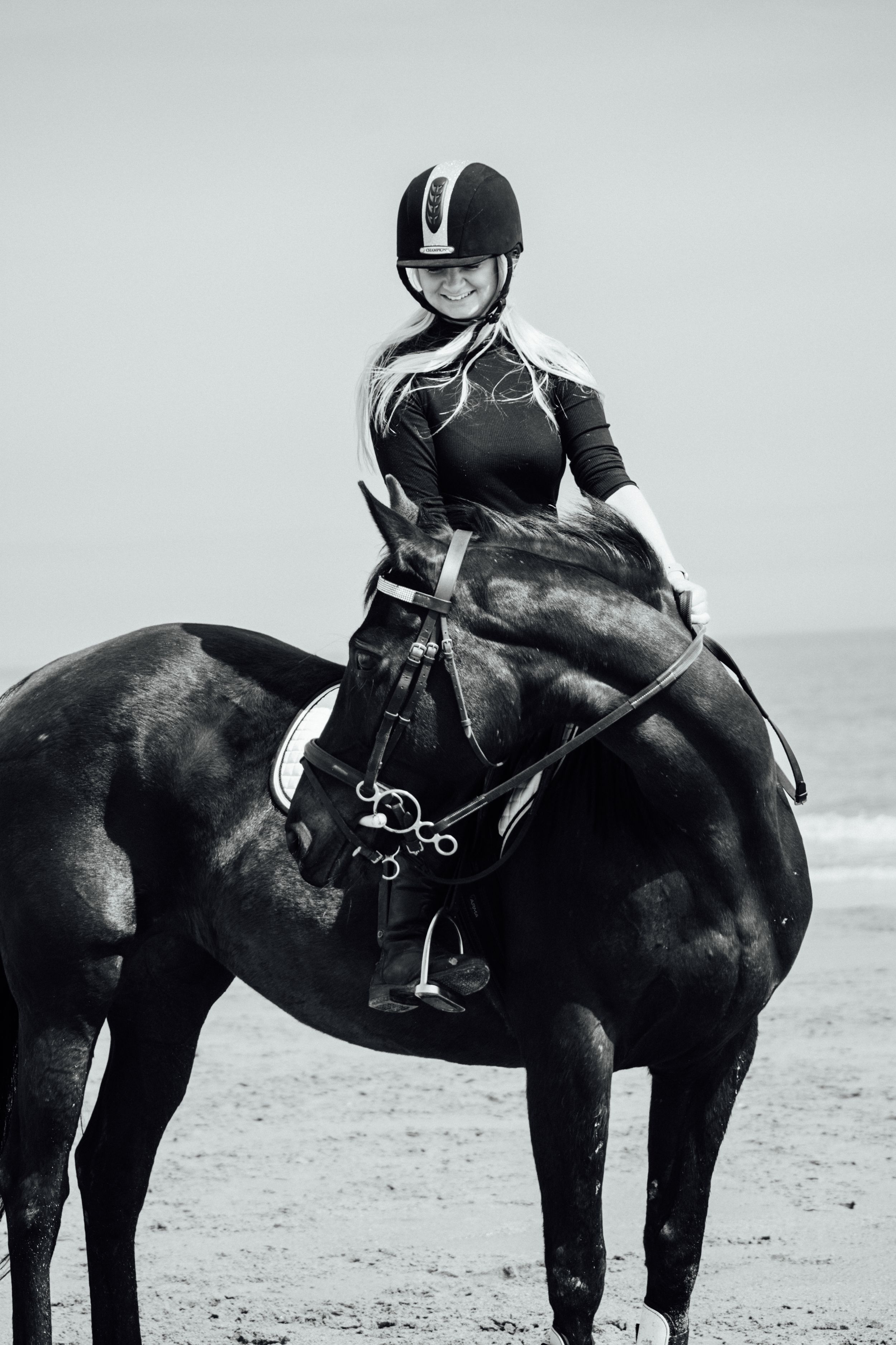 An equestrian in professional riding attire sits mounted on a black horse at the beach.