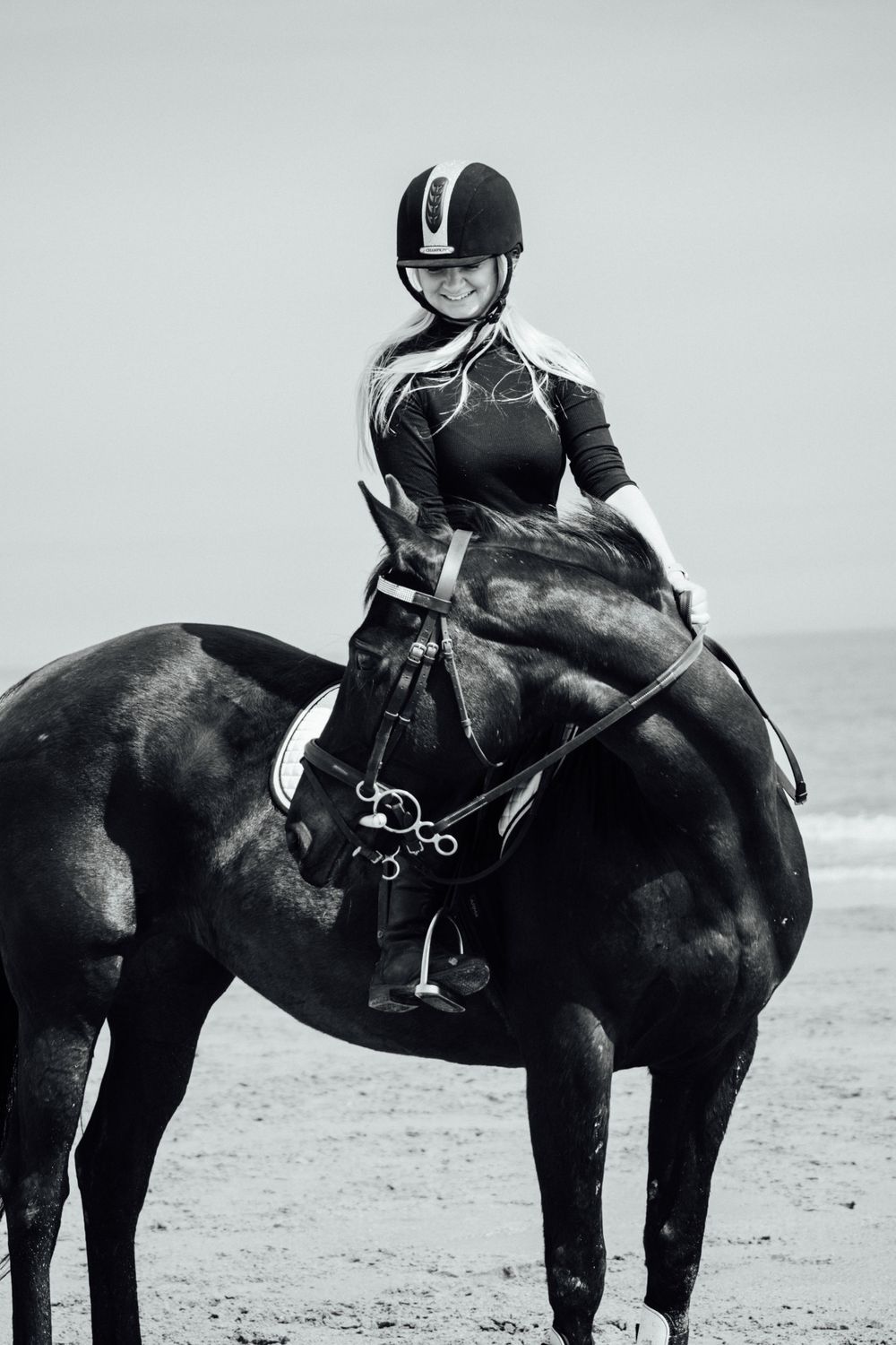 An equestrian in professional riding attire sits mounted on a black horse at the beach.