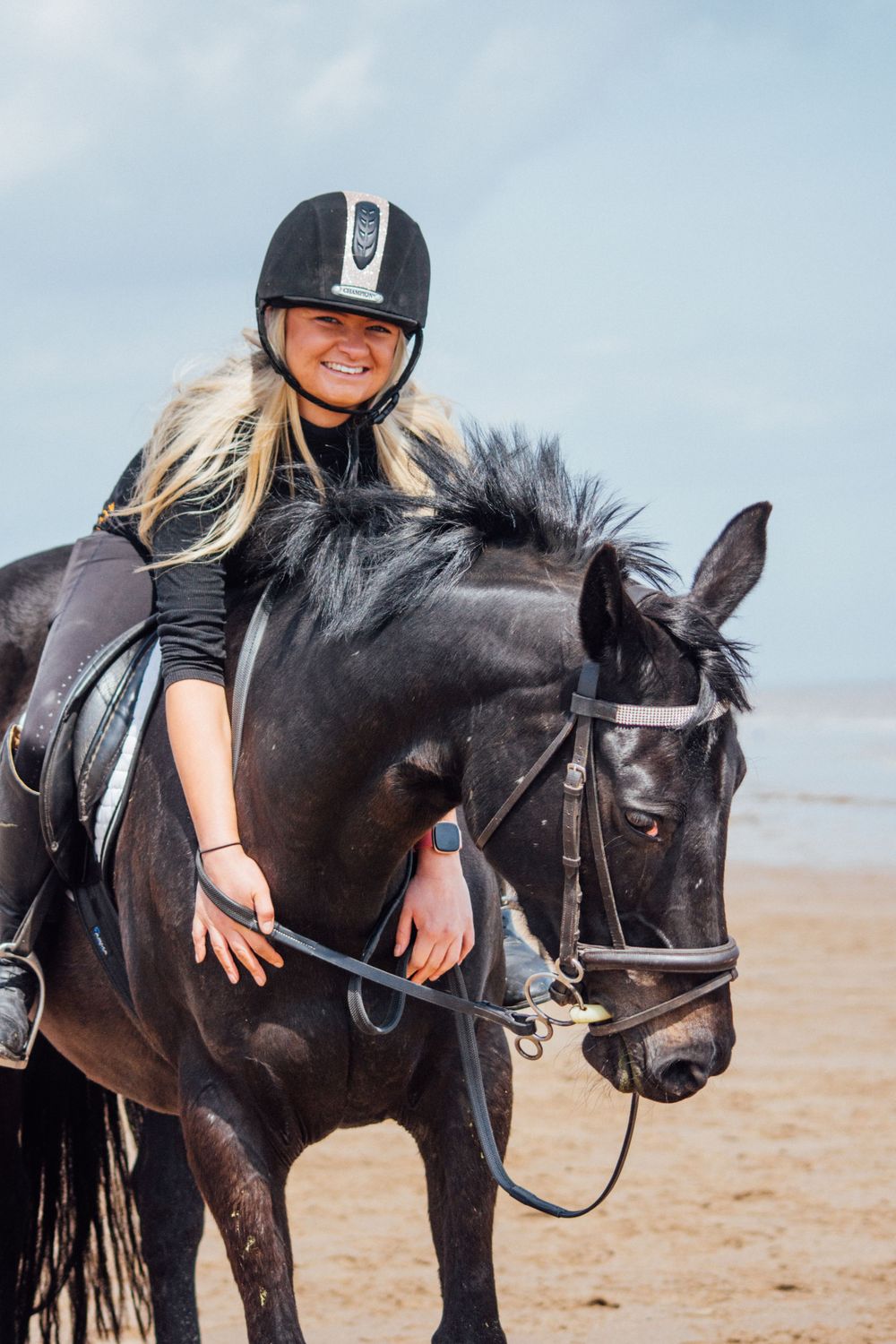 A mounted rider in protective gear sits atop a black horse on the beach.