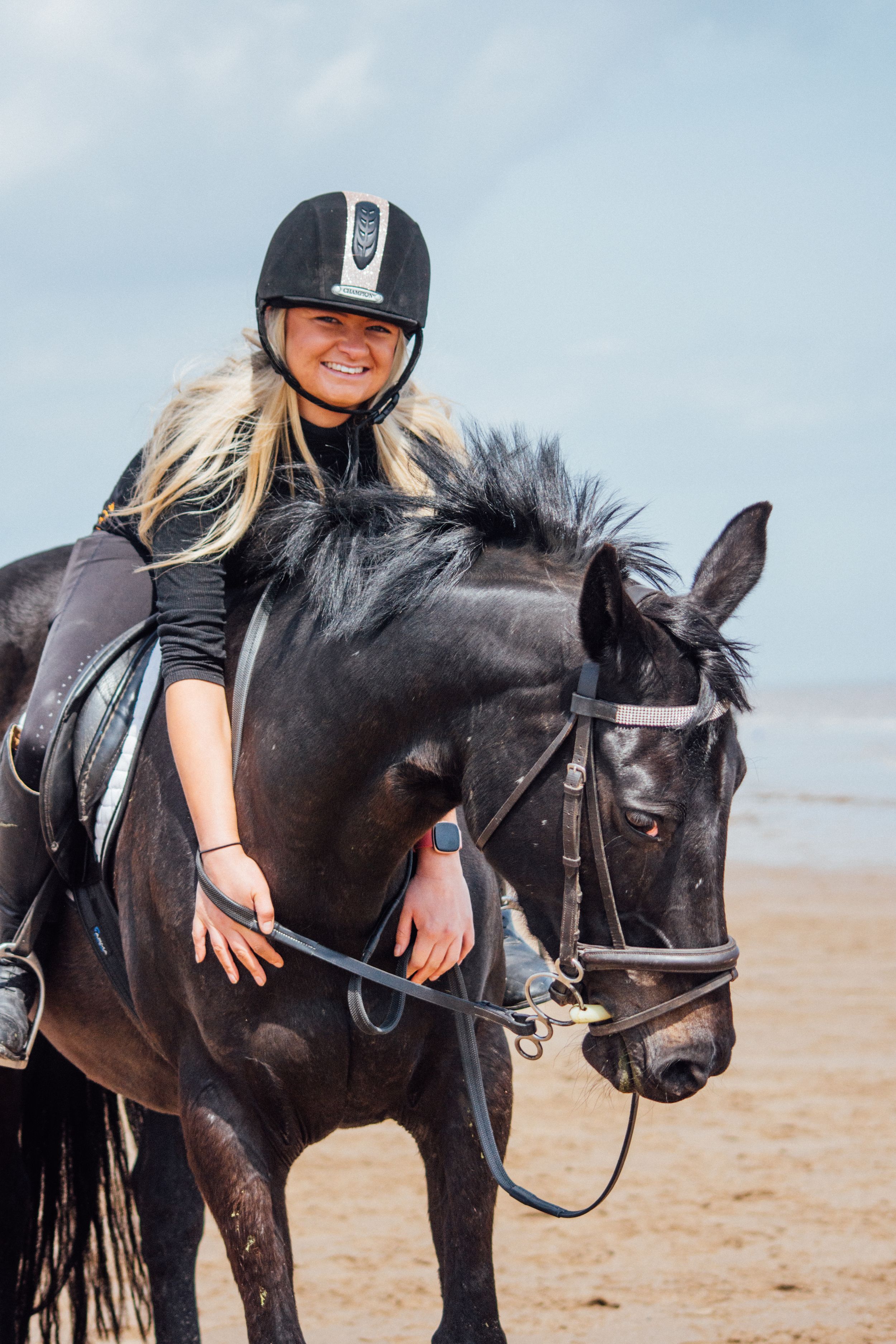 A mounted rider in protective gear sits atop a black horse on the beach.
