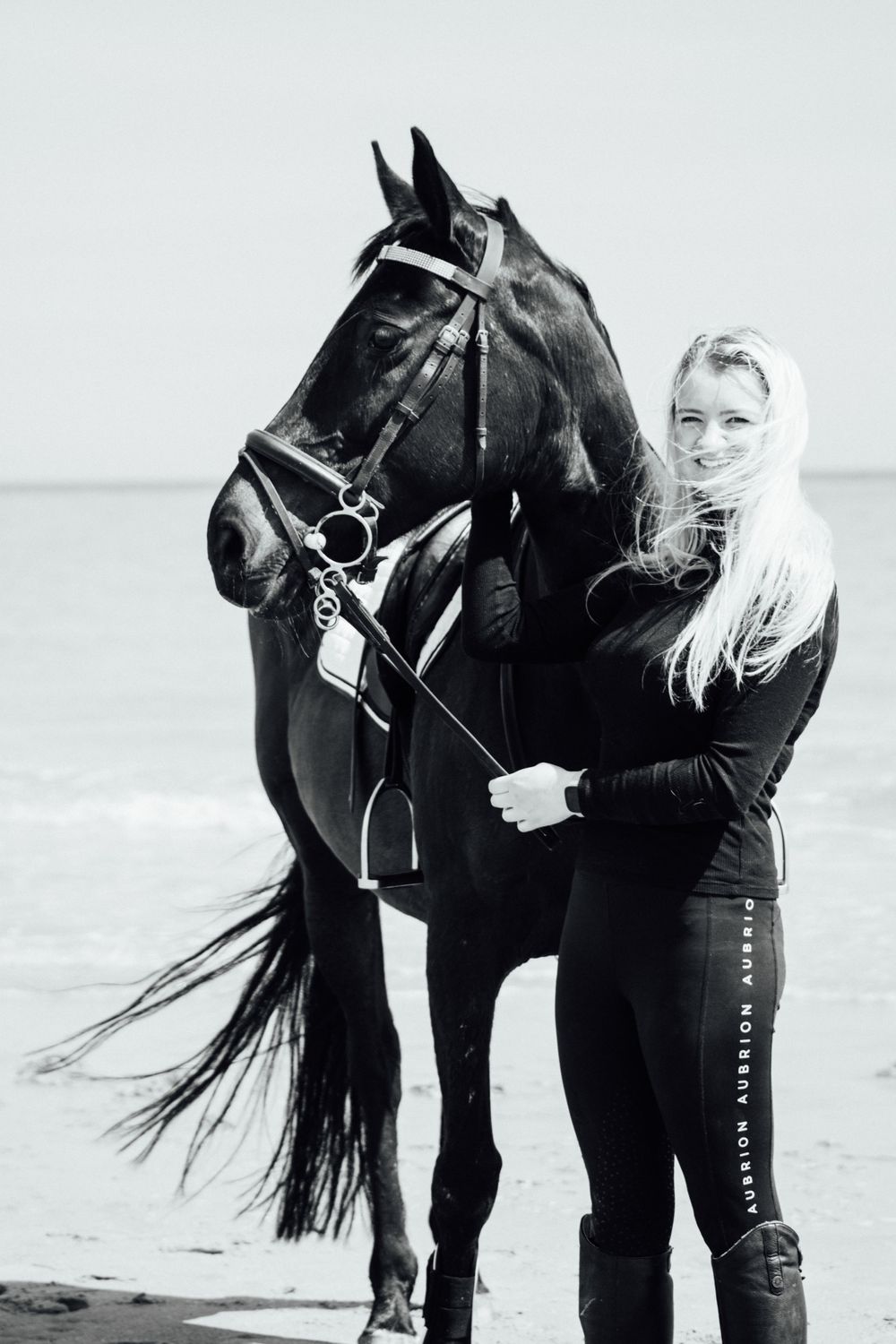 A striking black horse with full tack stands against an overcast beach backdrop.