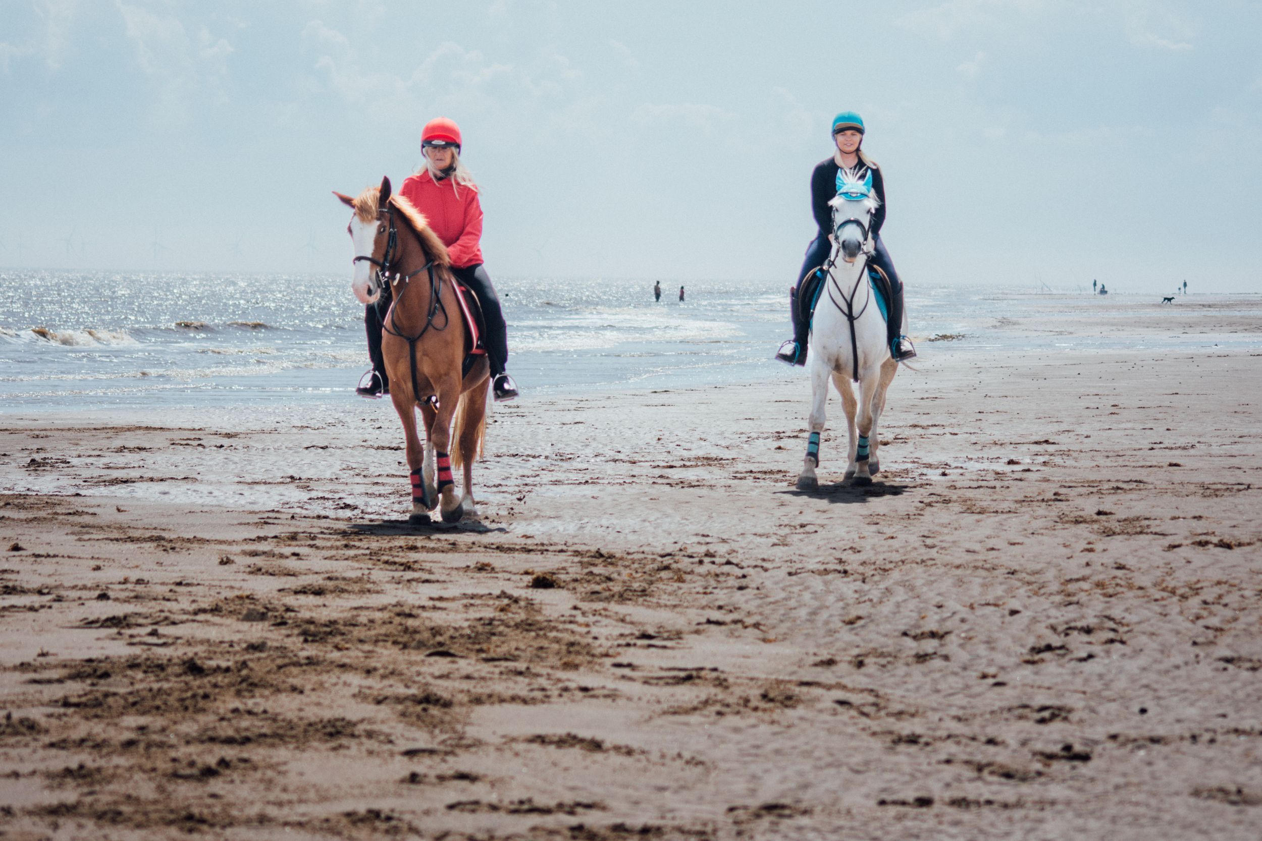 Two riders on horseback walking along a sandy beach with the ocean in the background.
