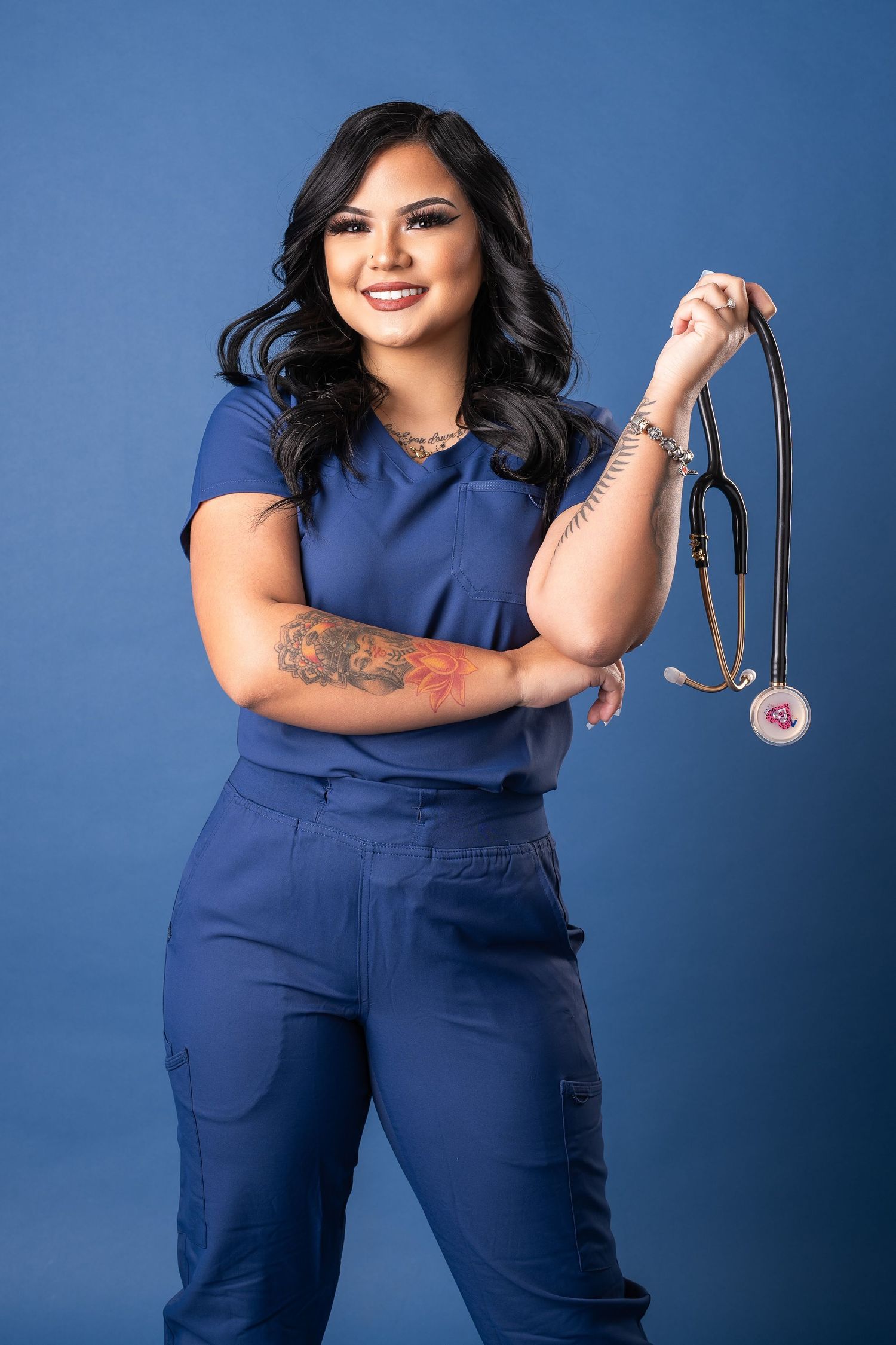 Healthcare worker in navy blue scrubs poses confidently against blue backdrop with stethoscope.