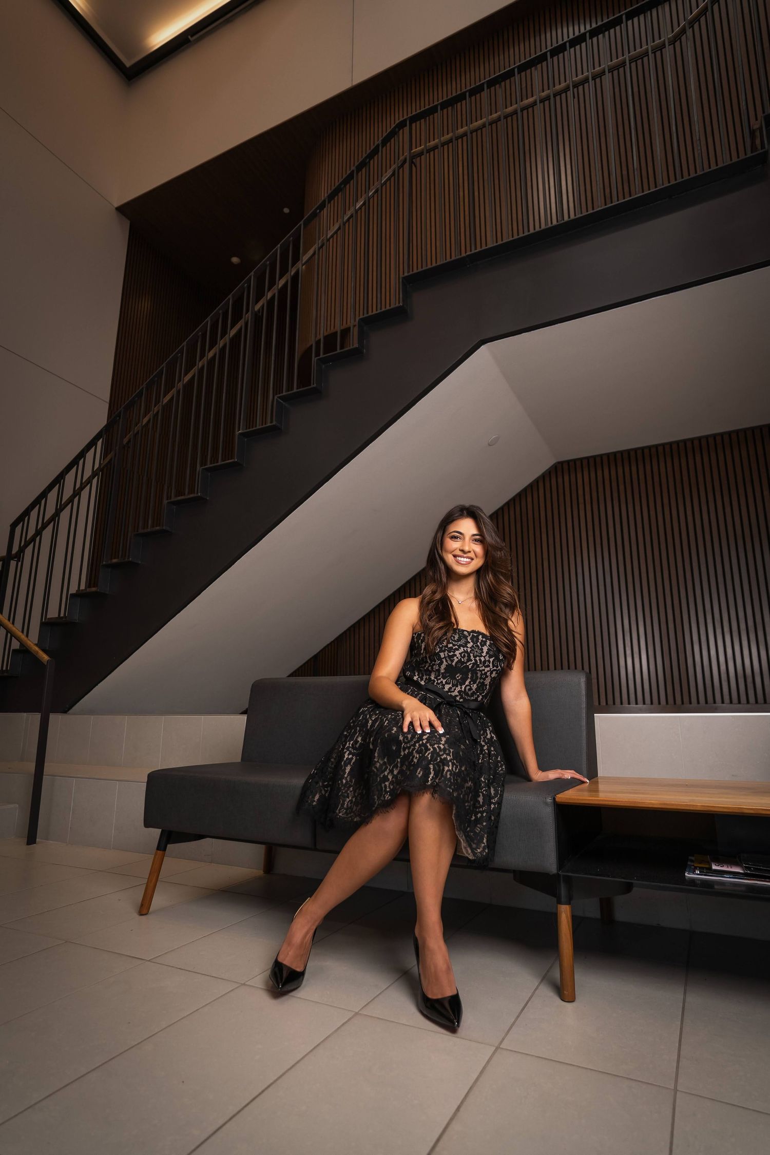 A person in a black dress poses elegantly on stairs in a modern interior setting with dramatic lighting.