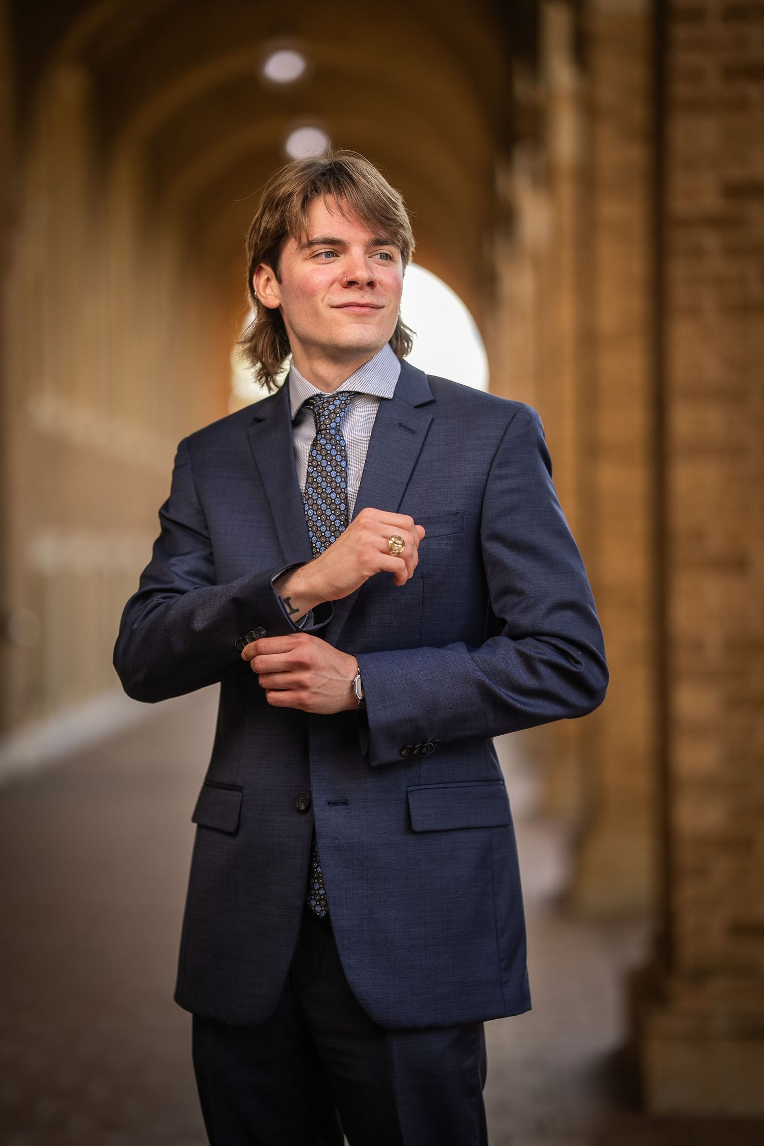 Young professional in navy suit and patterned tie adjusts cufflink while standing in arched stone hallway.