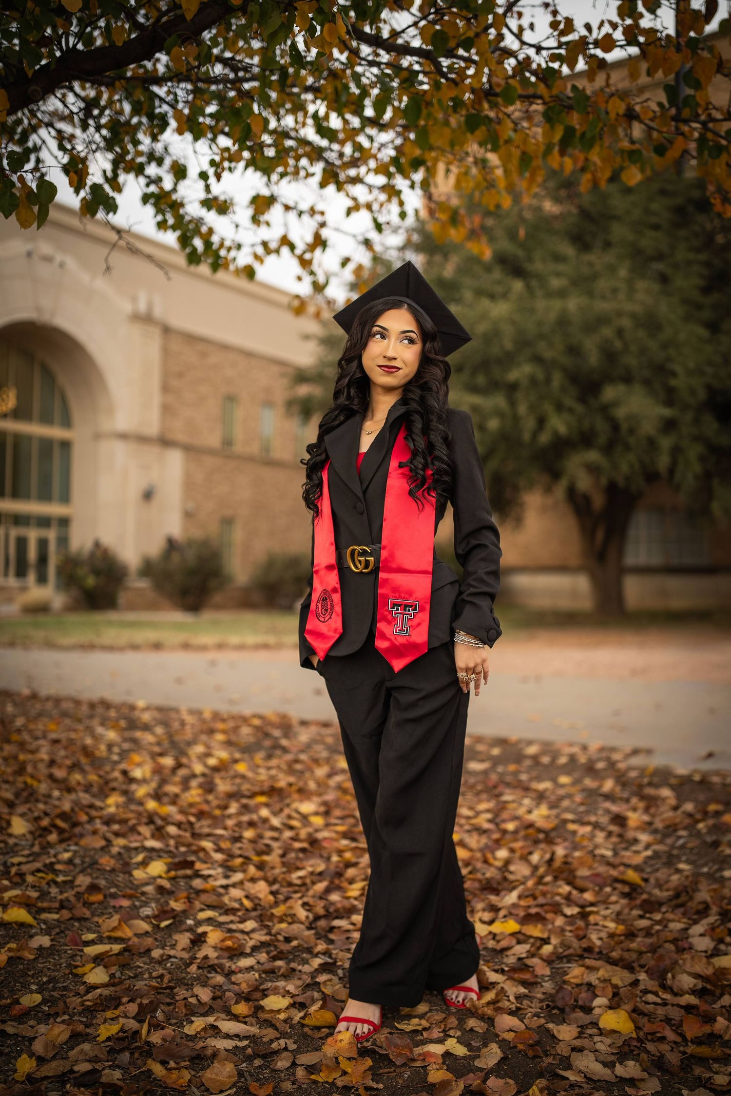 A person in graduation attire poses in autumn leaves wearing a black gown and red stole against a brick building backdrop.