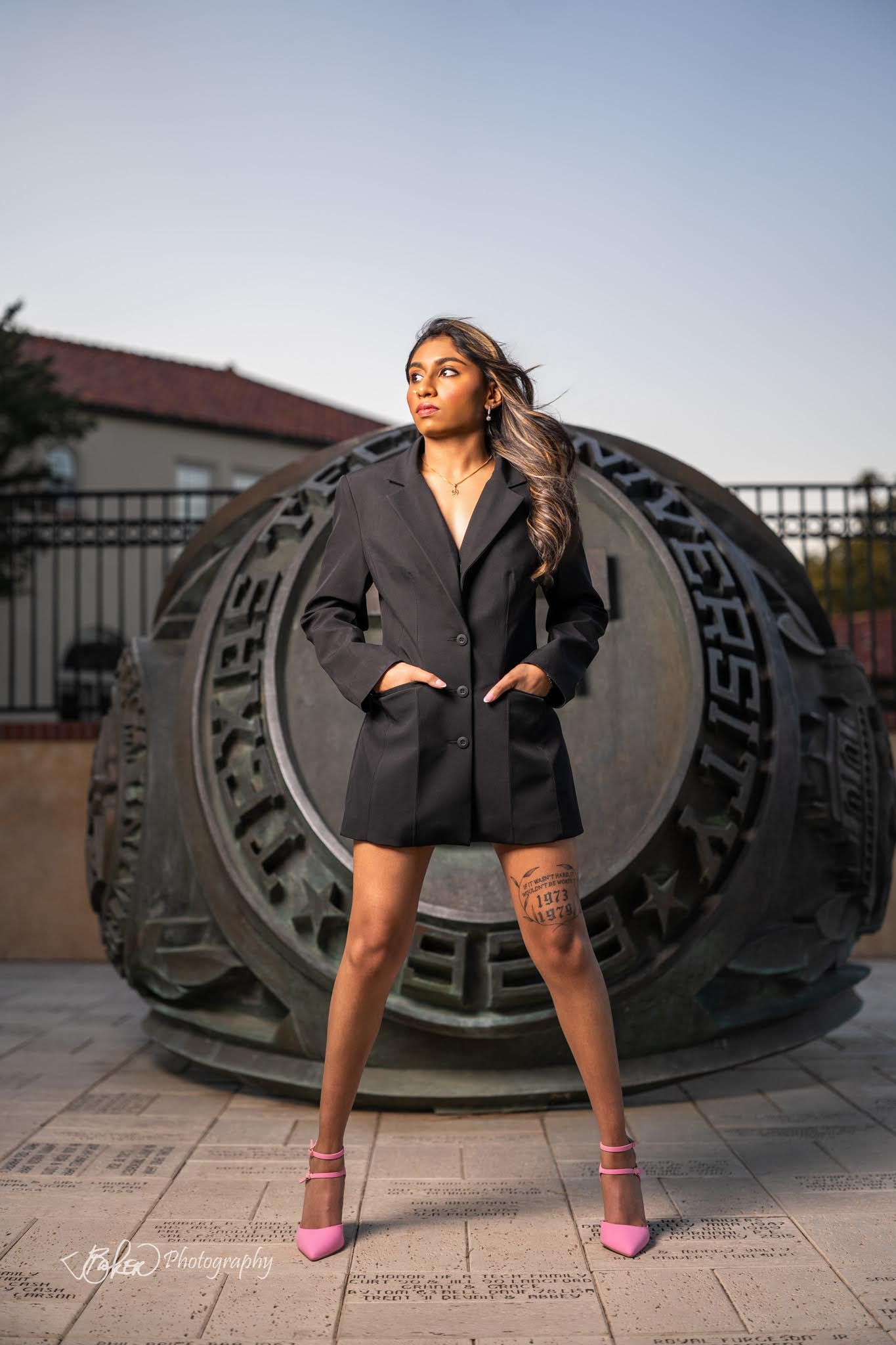 Fashion model in black blazer dress poses against large industrial metal backdrop in urban setting at sunset.