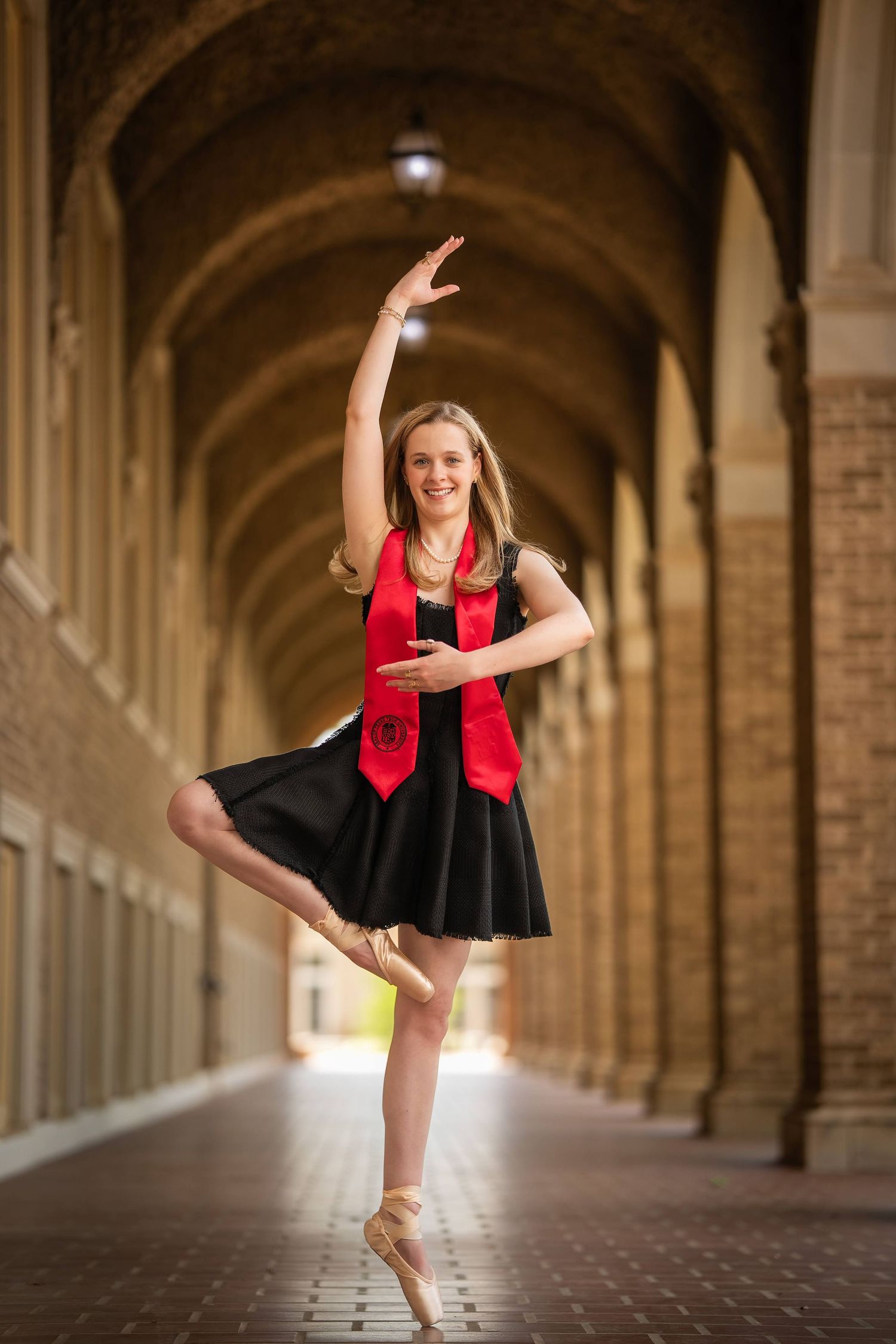 Dancer in black dress and red vest performs graceful high kick in arched stone hallway.