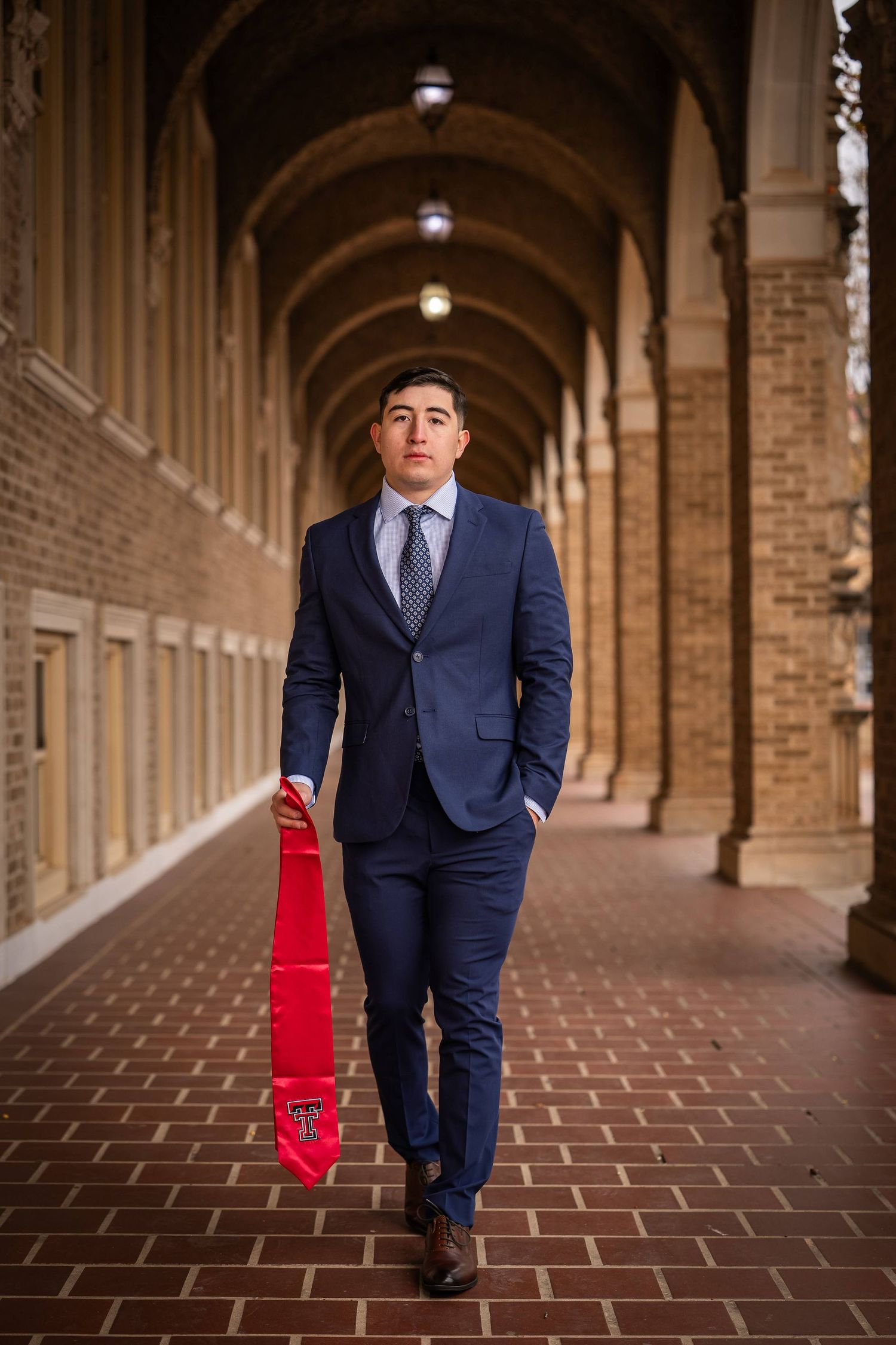 A person in a navy suit walks along a brick archway corridor holding a red Texas Tech banner.