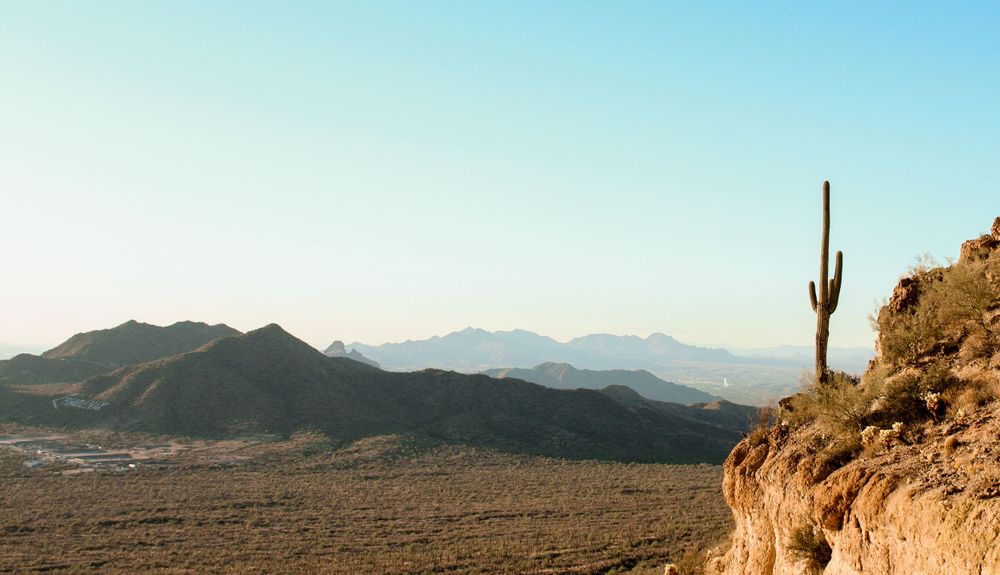 Lone saguaro cactus on rocky cliff overlooking vast desert valley with mountain range at sunset Arizona.