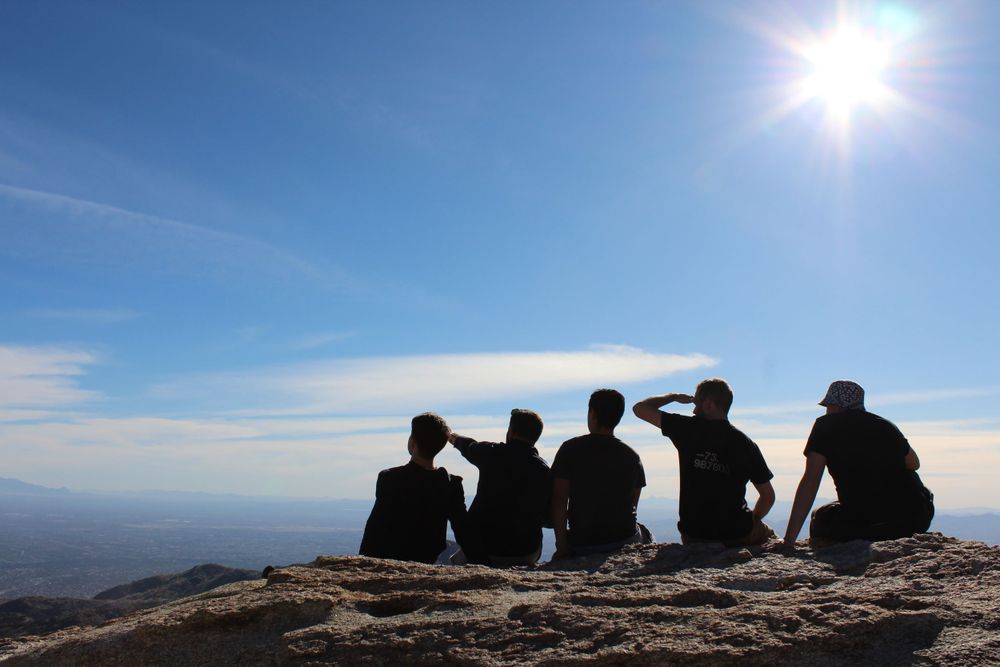 Group of friends silhouetted against blue sky sitting on mountain summit enjoying panoramic views.