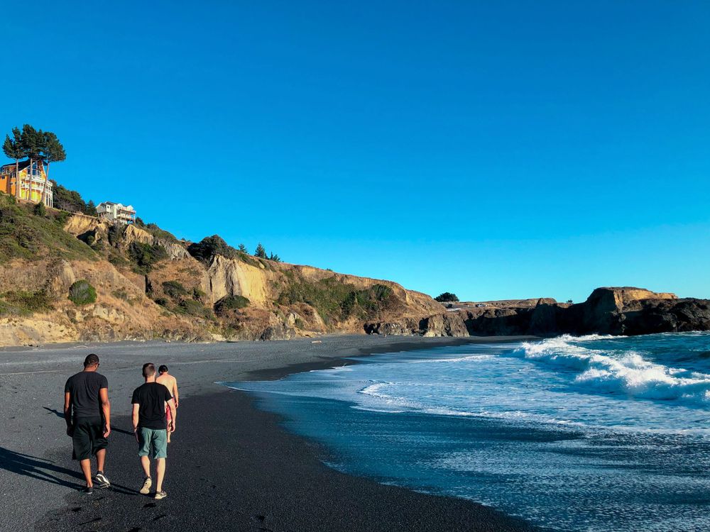 Two people walking along a black sand beach with crashing waves and coastal cliffs under clear blue sky.