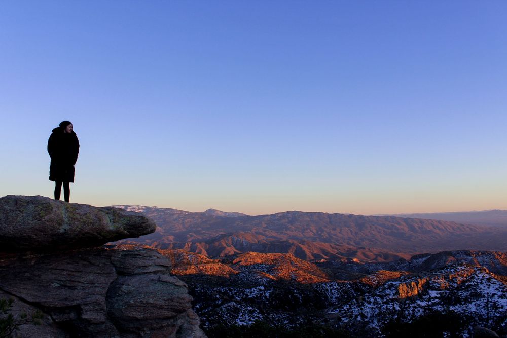 Silhouette of a person standing on a rocky mountain peak overlooking a vast snow-dusted canyon landscape at sunset.