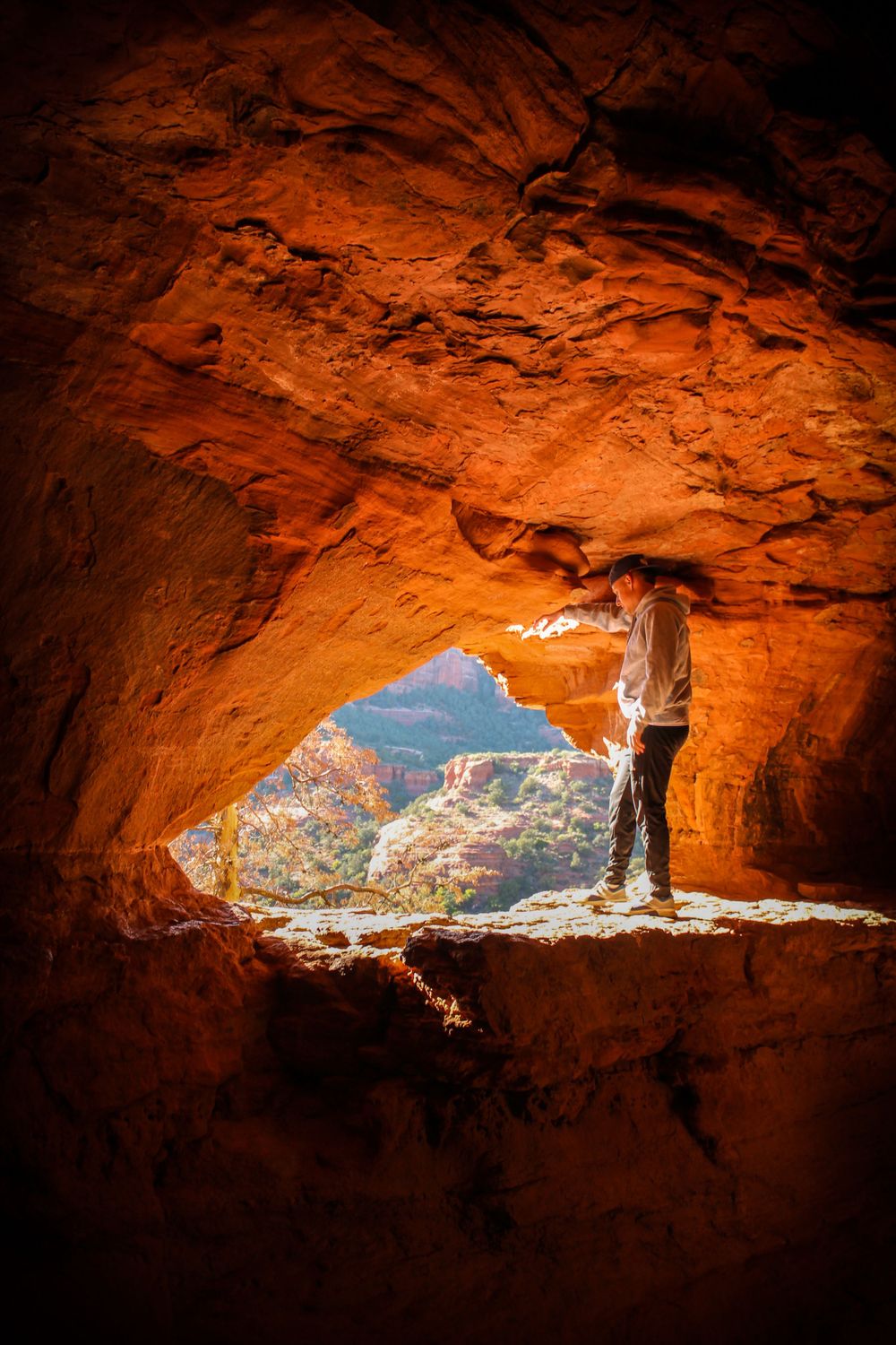 Hiker standing in a glowing red sandstone cave opening overlooking a scenic canyon valley in Sedona, Arizona.