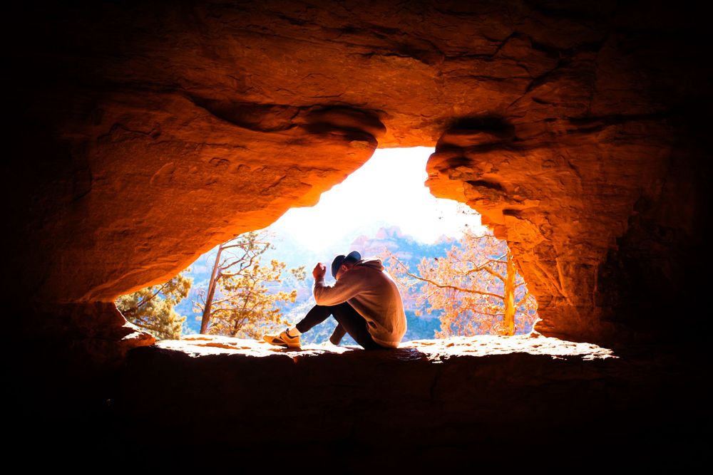 Person sitting alone in a red sandstone cave entrance with colorful autumn foliage visible through the opening.