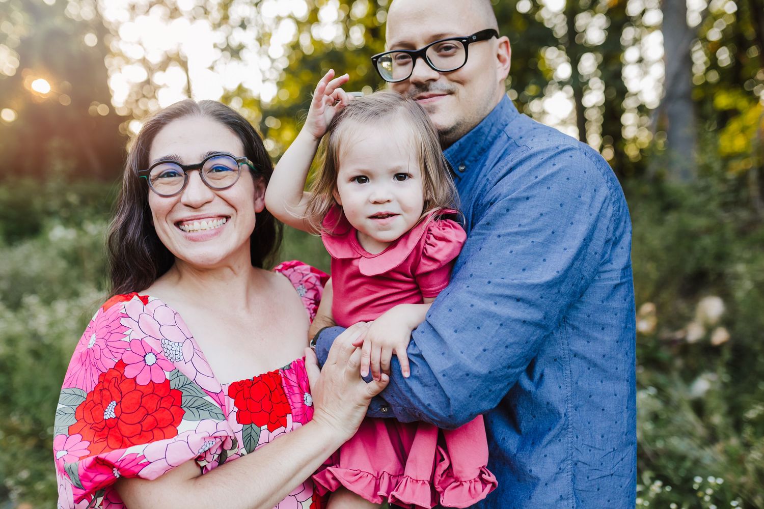 Mom wearing pink and red floral dress stands next to husband wearing blue and daughter in a pink dress