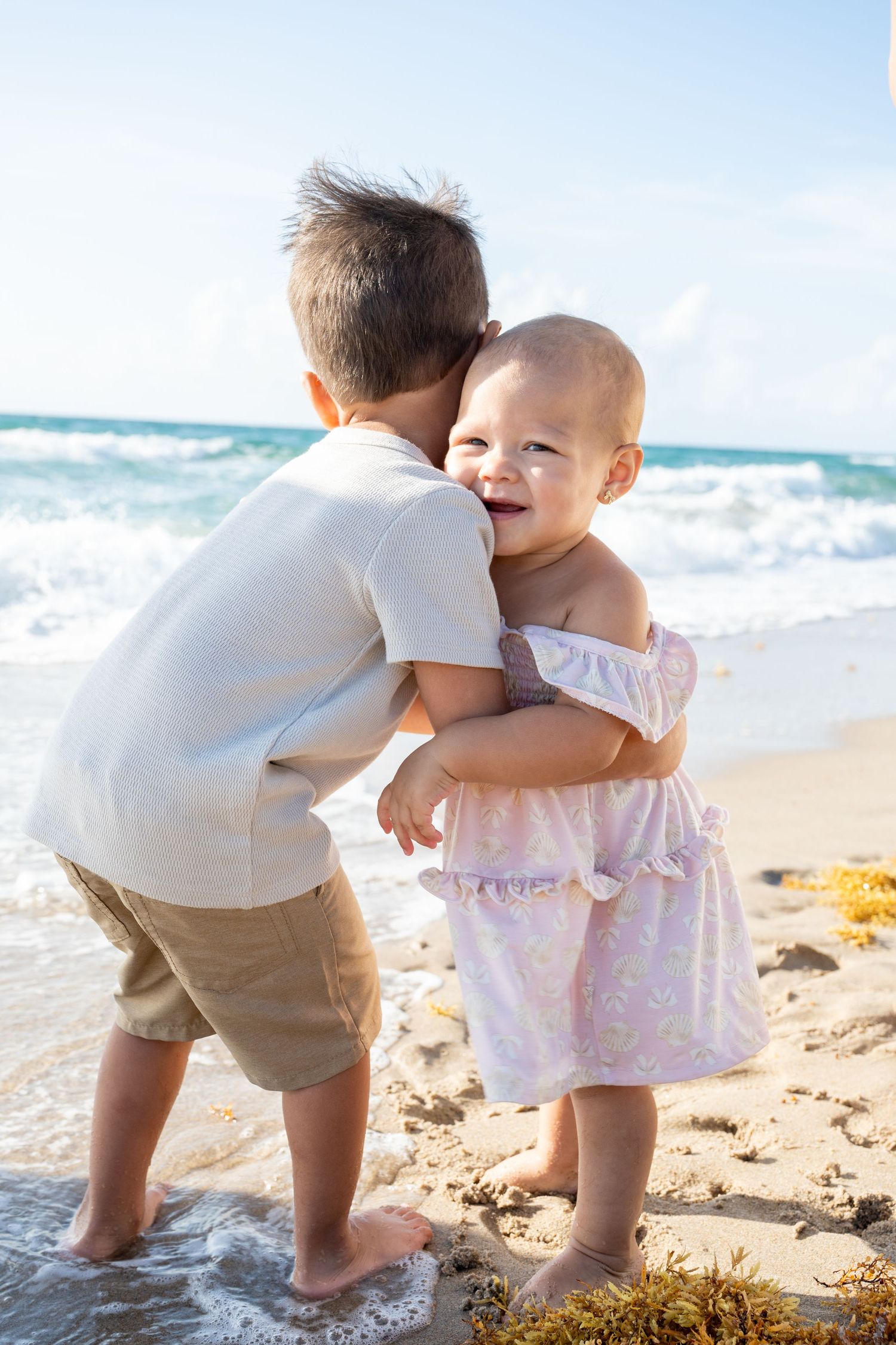 A toddler and adult play together on a sunny beach with waves crashing in the background.