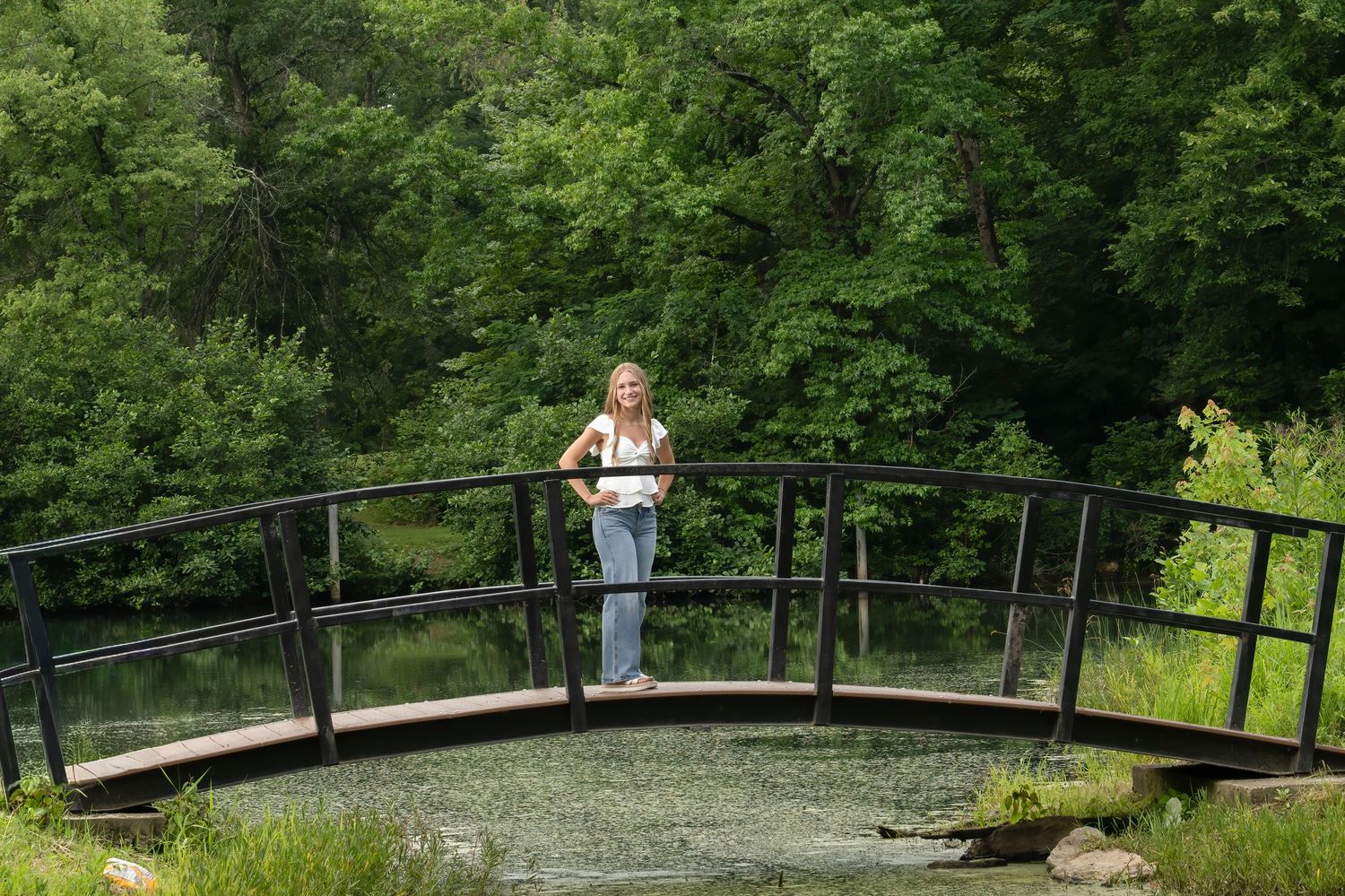 Person sitting by a creek in a wooded area wearing casual clothes and white sneakers against a natural forest backdrop.