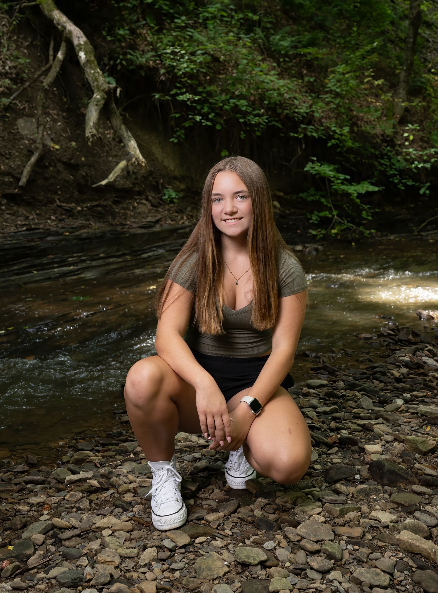 Person sitting by a creek in a wooded area wearing casual clothes and white sneakers against a natural forest backdrop.