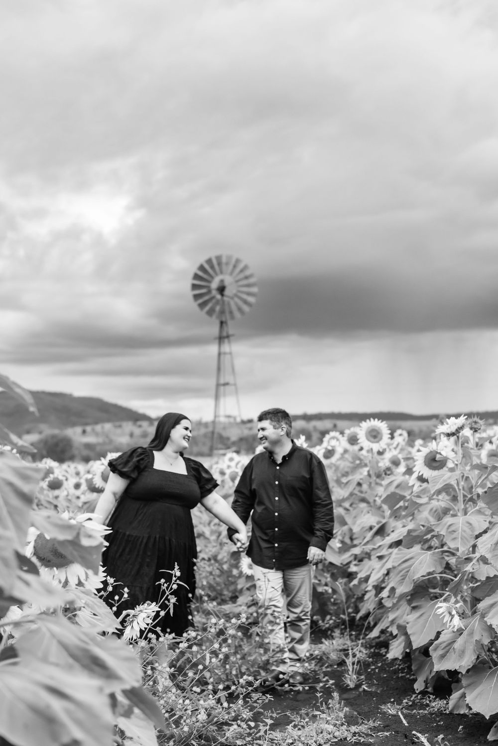 Romantic couple standing in sunflower field with vintage windmill and moody sky in black and white photography.