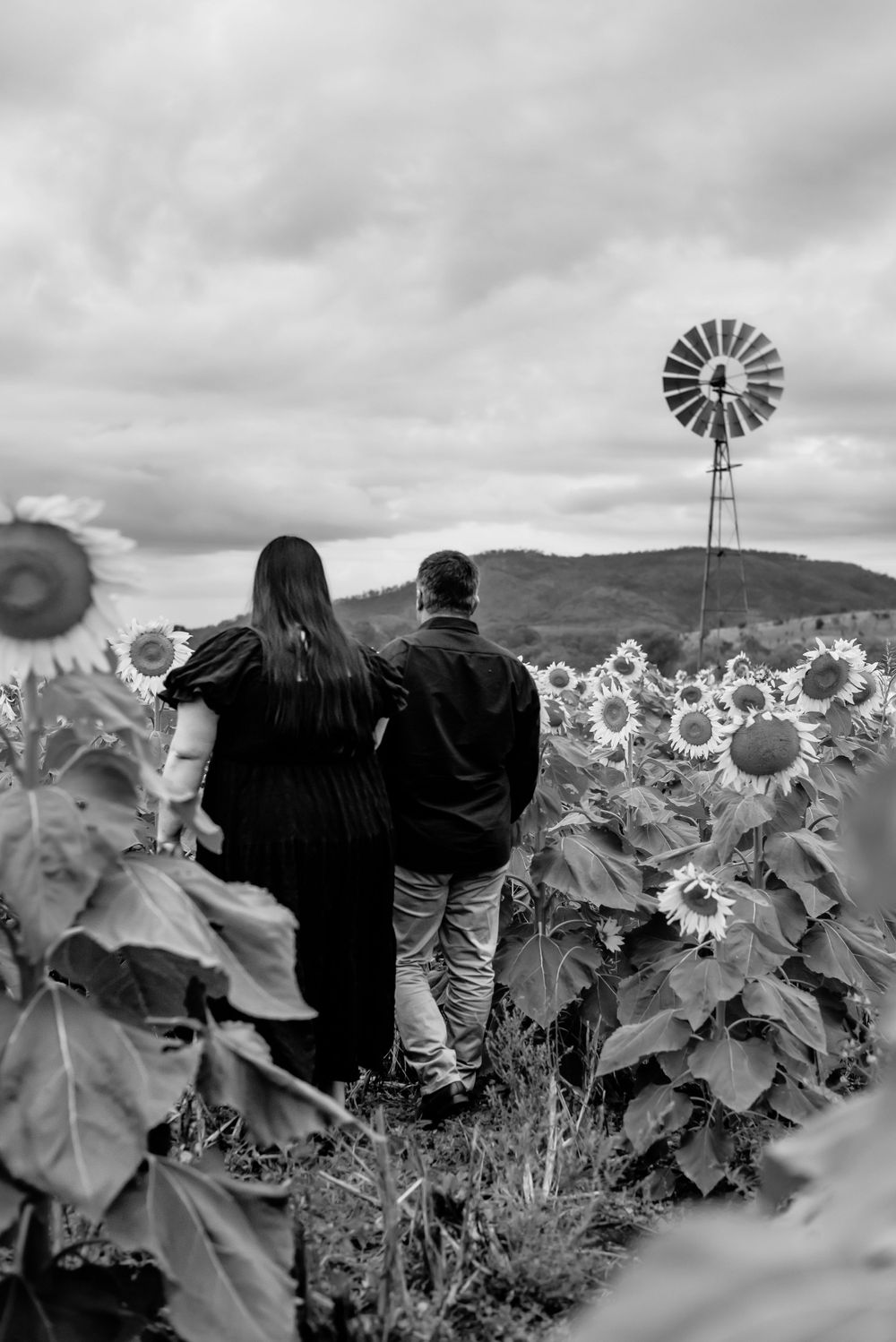 Couple embracing while viewing windmill in sunflower field under cloudy sky.