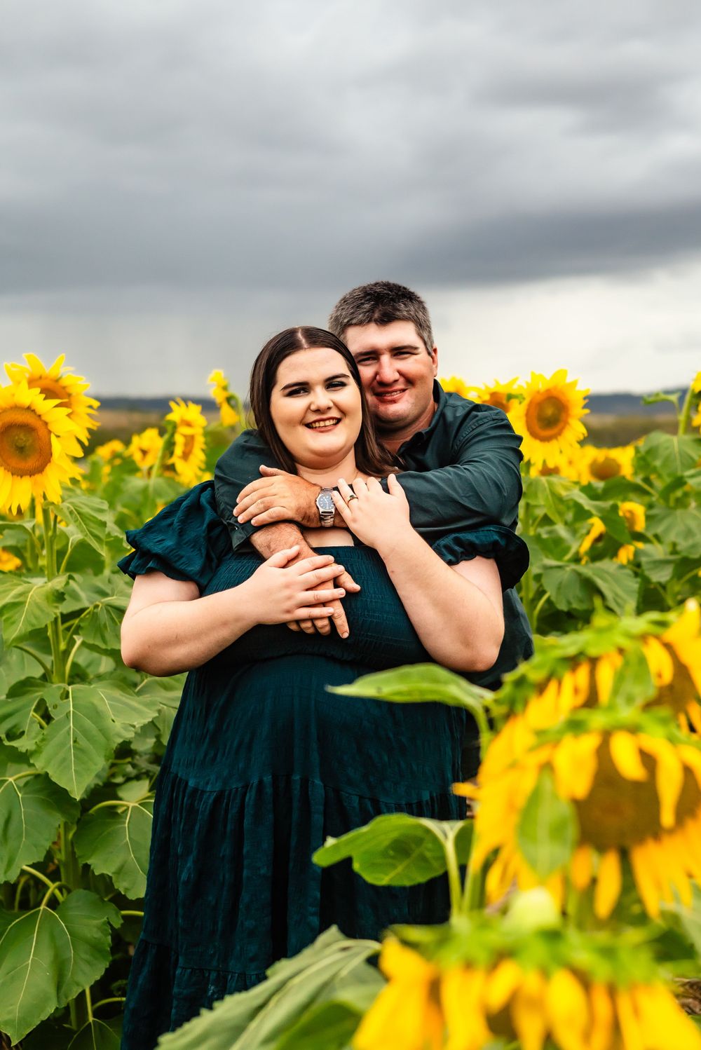 Happy couple embracing in colorful sunflower field with dramatic clouds overhead.