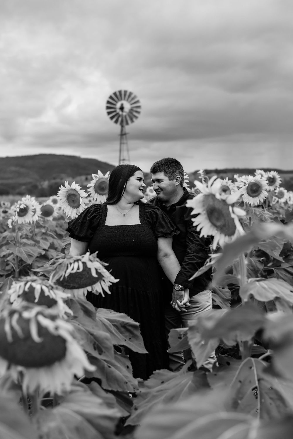 Couple sharing tender moment in black and white photo amid tall sunflowers and rustic windmill.
