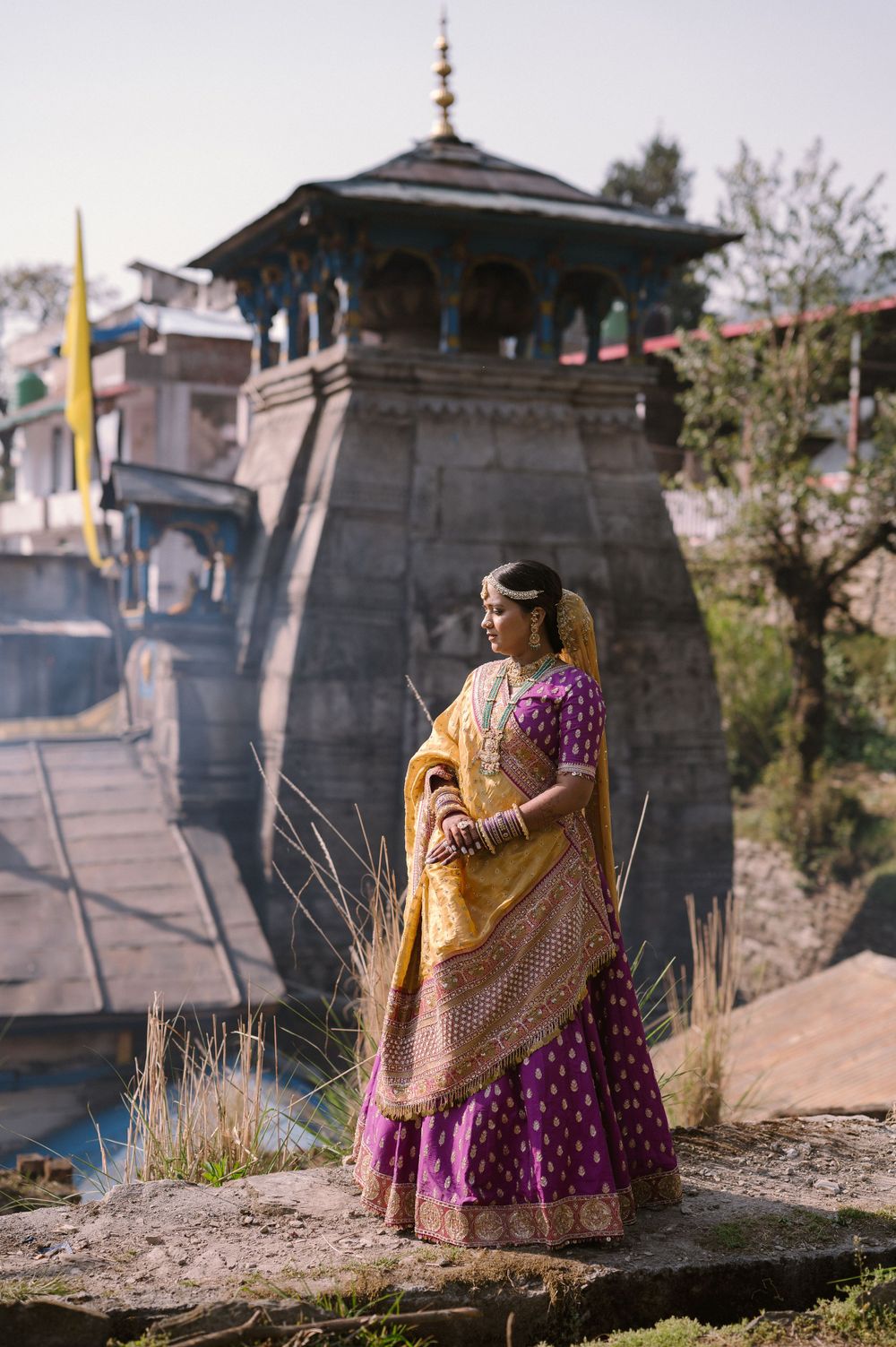 Traditional Indian bride in purple and gold lehenga poses against ancient temple architecture.