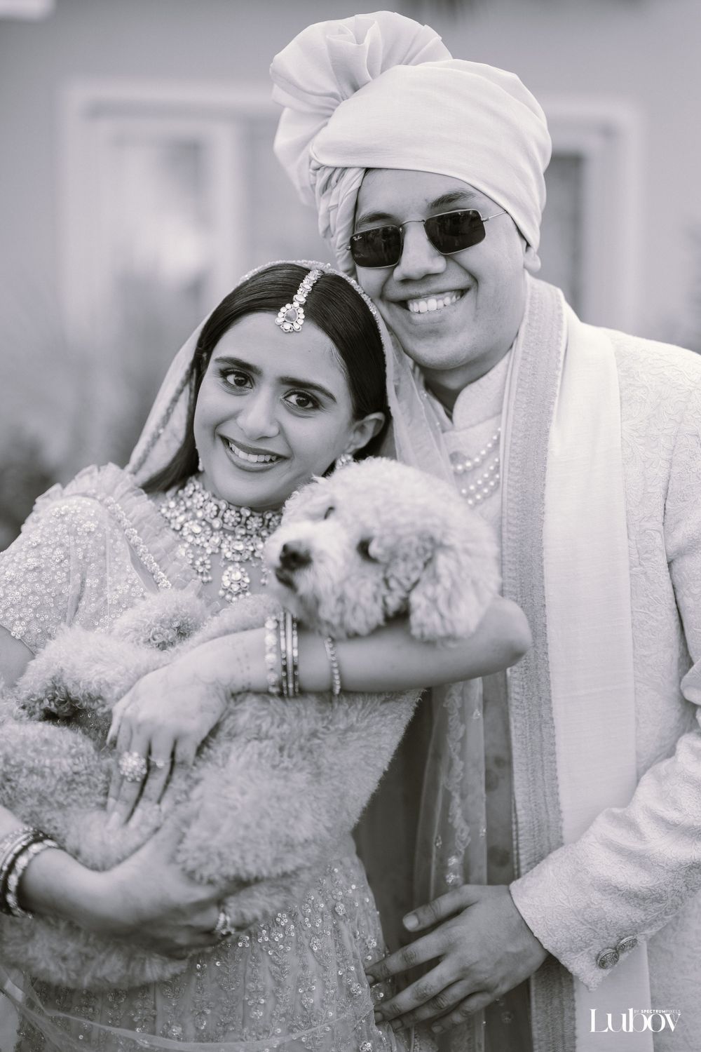 Couple in white formal attire poses with their fluffy white dog in black and white photo.