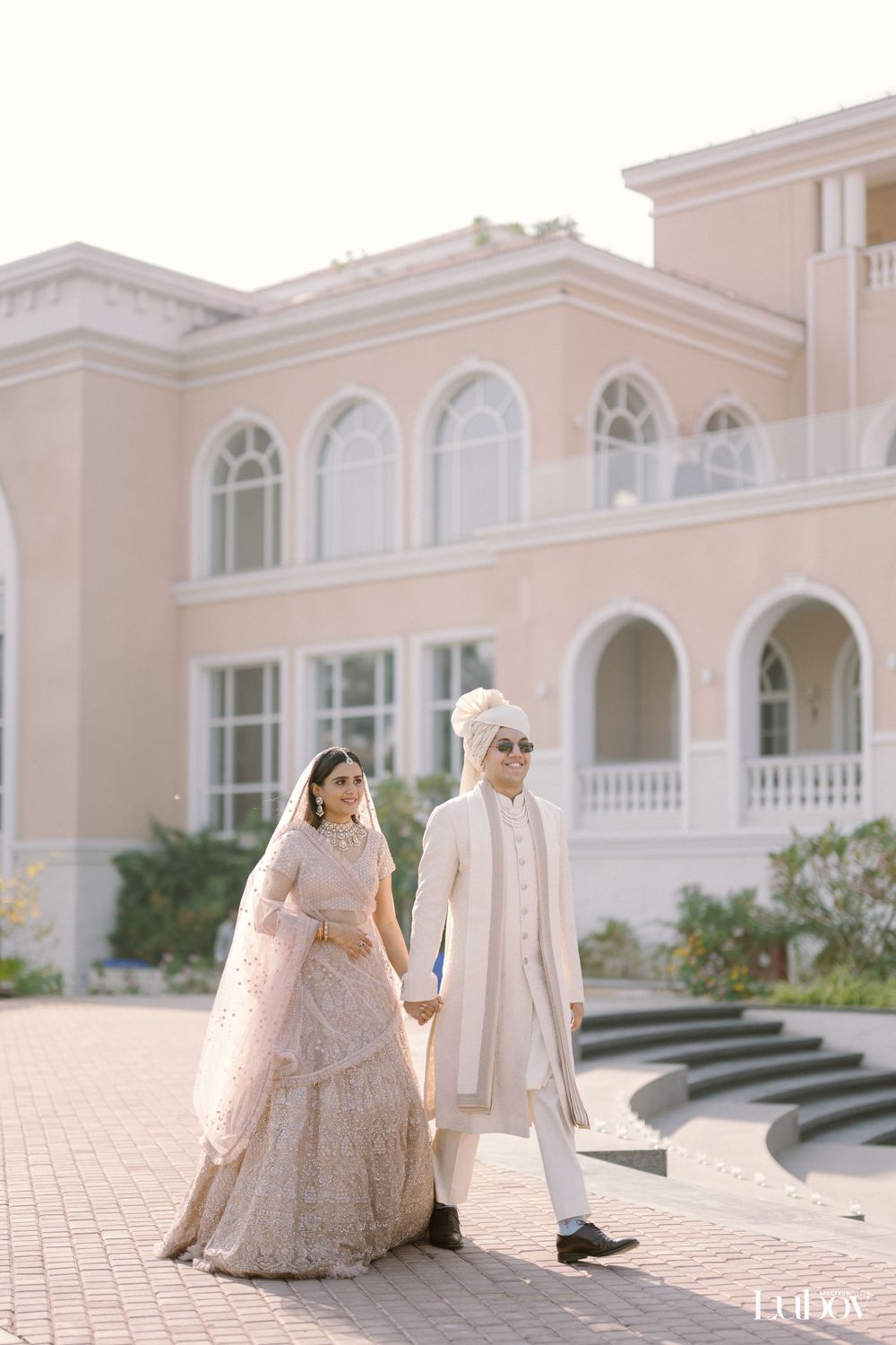 Couple in traditional wedding attire poses outside elegant pink Mediterranean-style building.