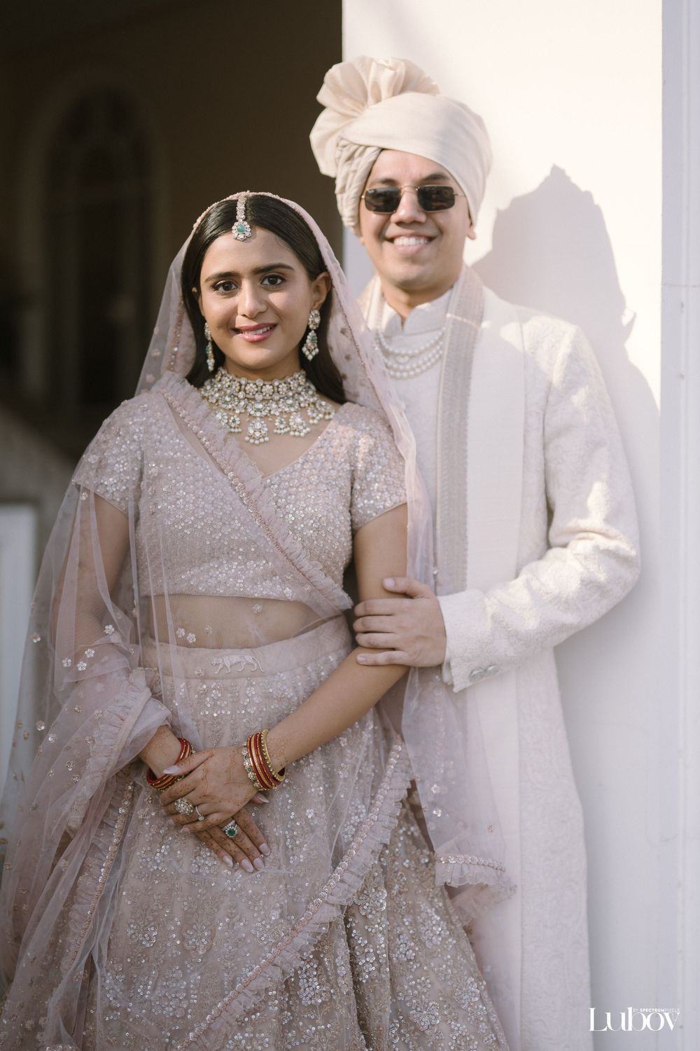 A couple poses in traditional Indian wedding attire with the bride in an embellished lehenga and groom in white sherwani.