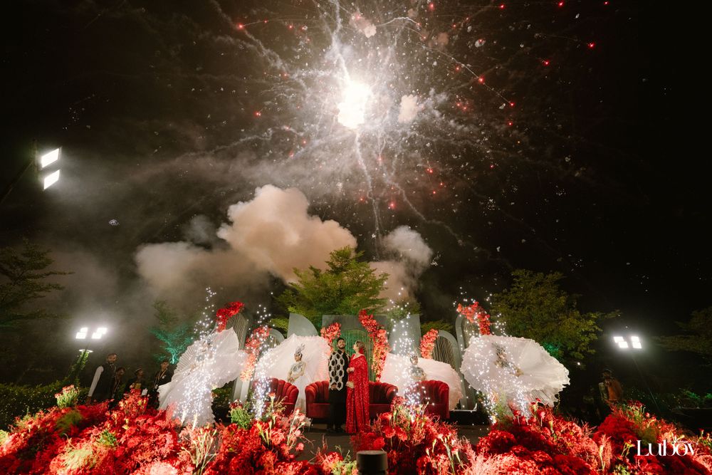 Spectacular fireworks display bursts over a wedding ceremony setup with red floral decorations and white canopies.