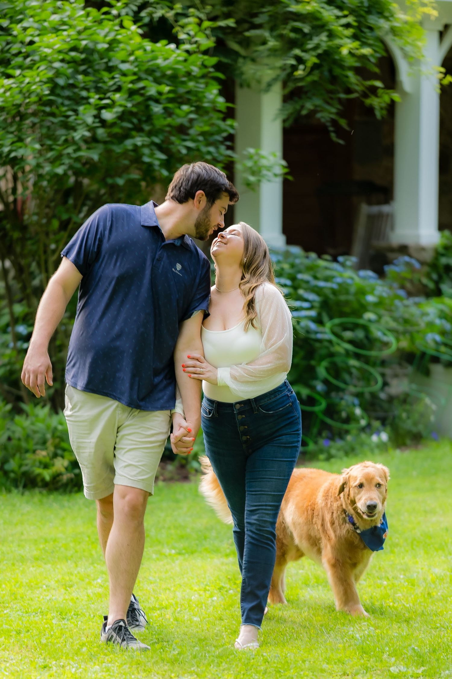 A couple and their golden retriever dog share a sweet moment while walking together in a lush green garden setting.