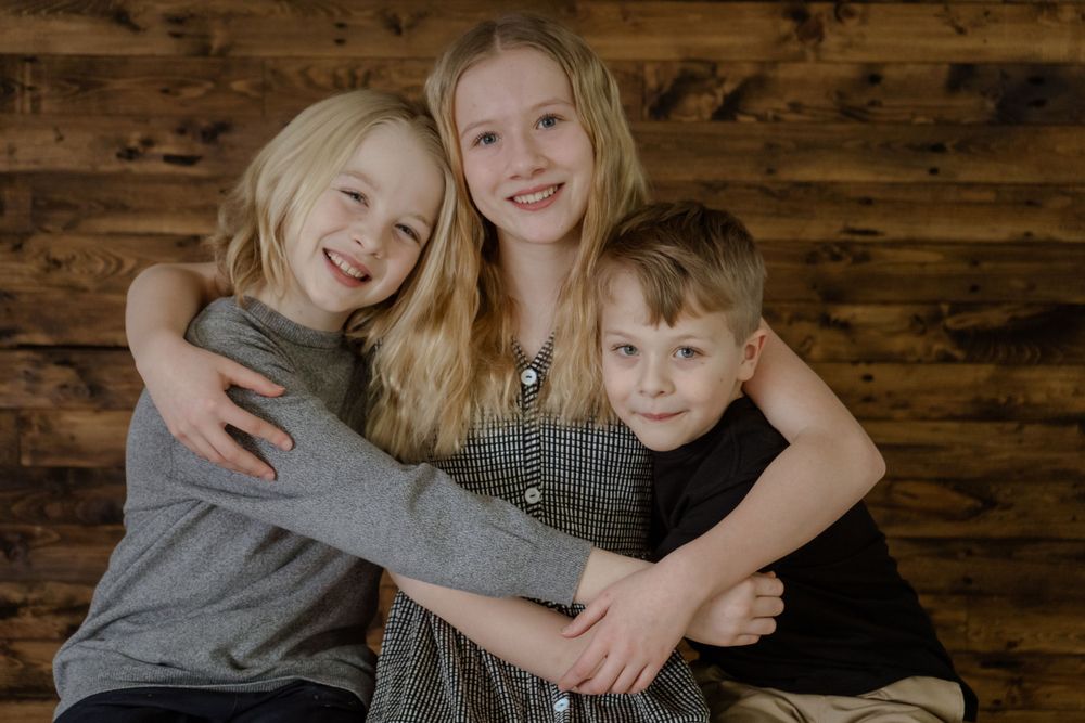 Three siblings sharing a warm embrace against a wooden wall background.