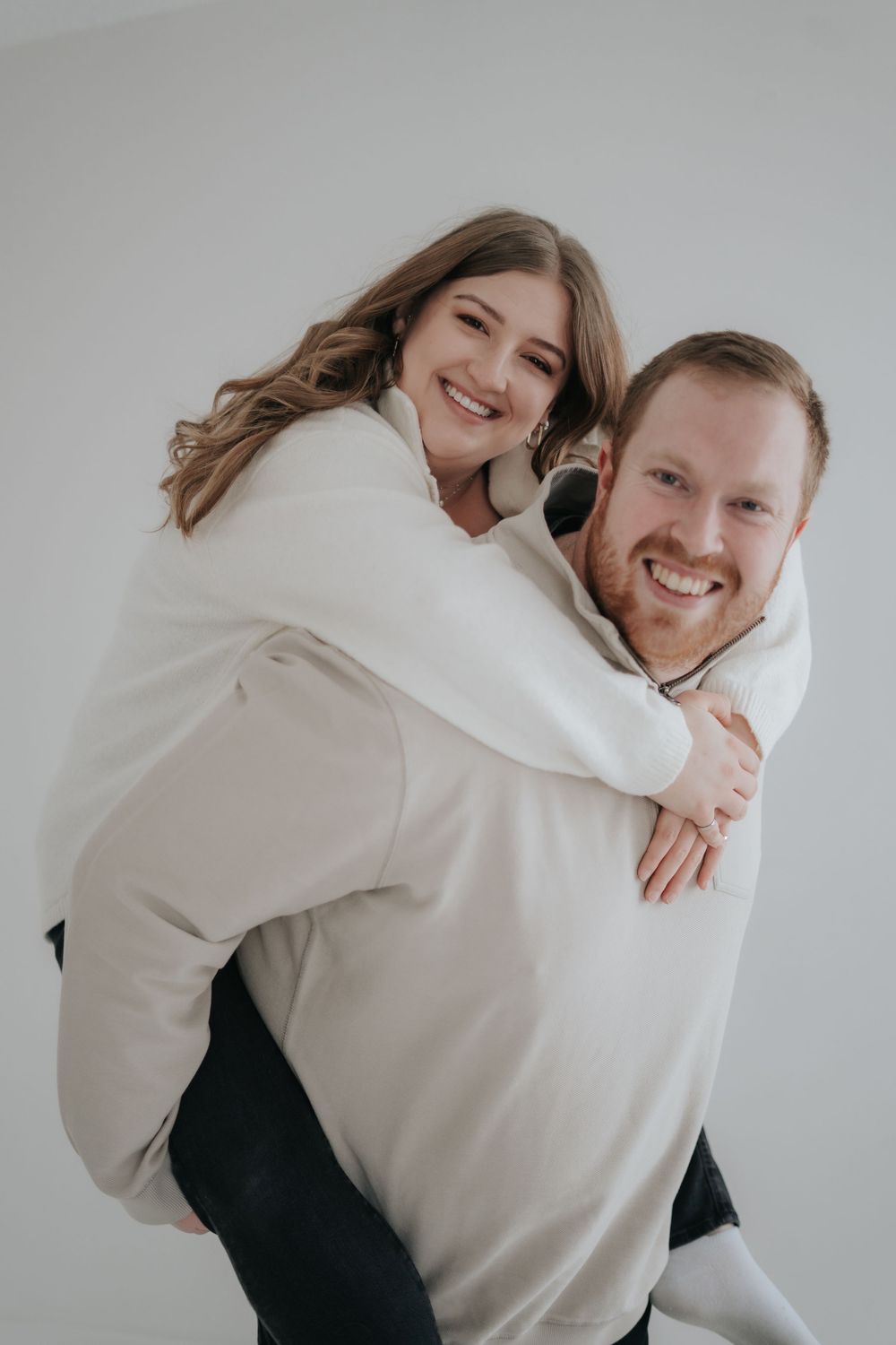 A couple wearing white sweaters share a playful piggyback pose against a light background.