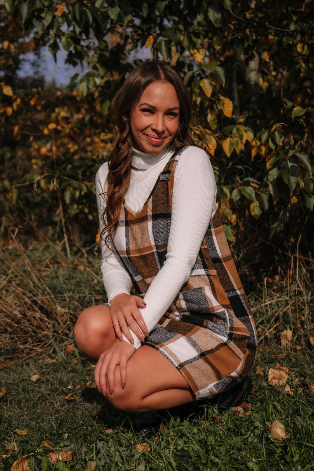 Person in white turtleneck and plaid vest sitting on grass during fall photo session.