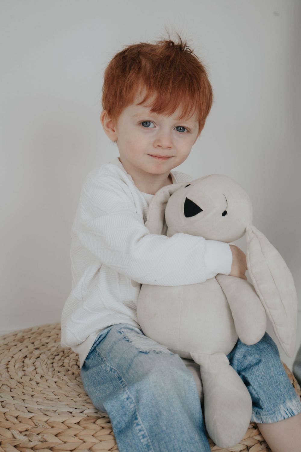 A young child in white sweater holding a plush teddy bear while sitting on woven ottoman.