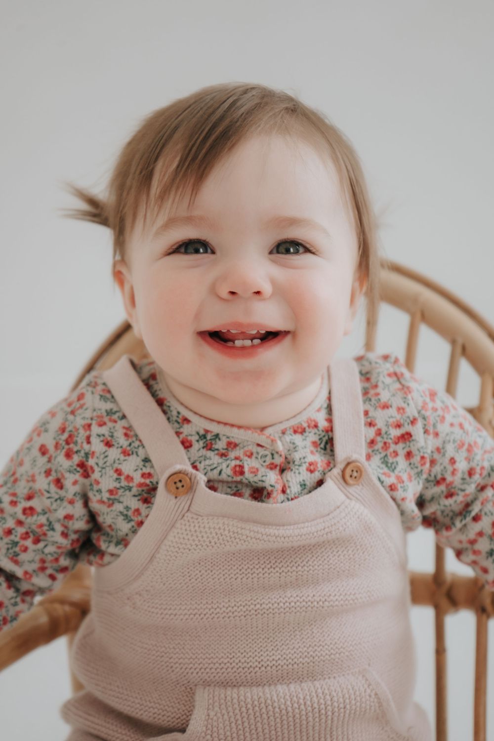 A happy toddler wearing floral print and beige overalls smiles brightly at the camera.